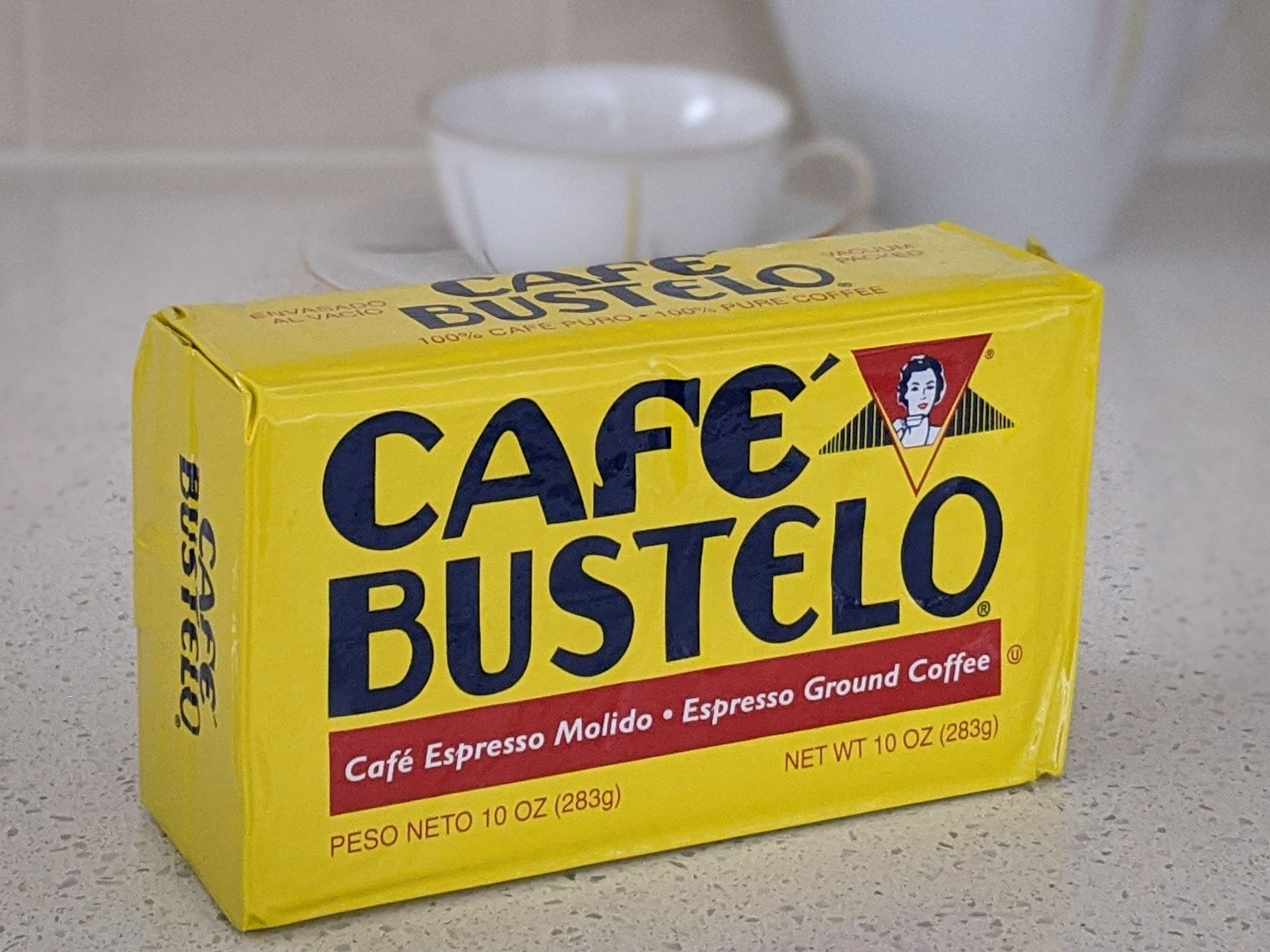 Café Bustelo Espresso Vacuum-Packed Dark Roast on a beige countertop, with a white porcelain kettle, tea cup, and saucer against a beige tile wall background