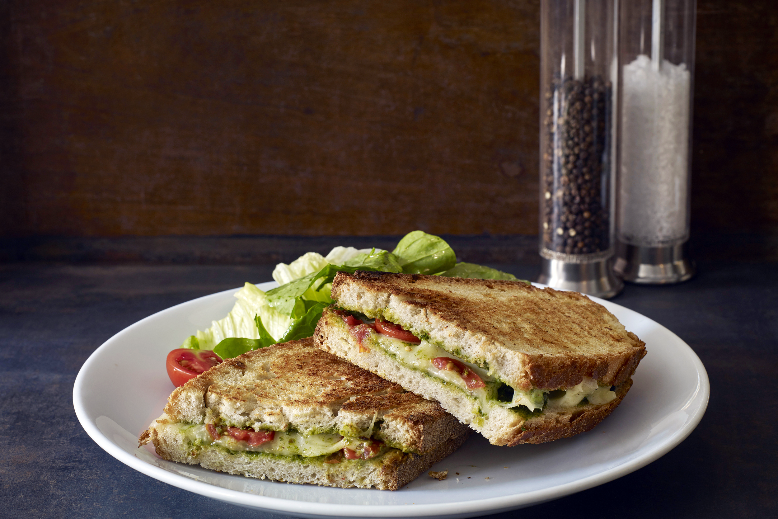 Caprese grilled cheese cut in half on a white porcelain plate, salt and pepper grinders to the right of it, on a blue tablecloth with a dark background