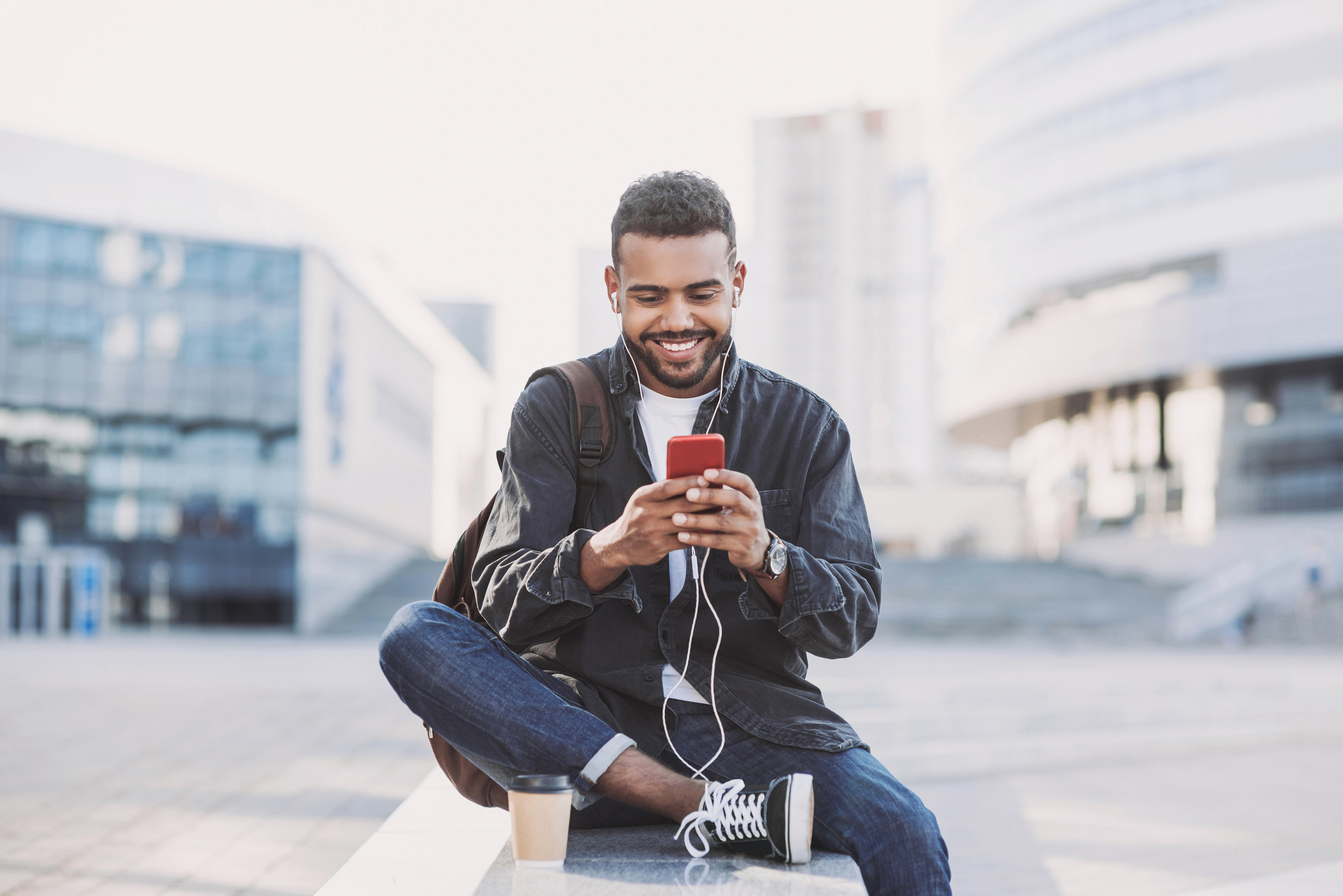 Cheerful young man using a smartphone while sitting on a ledge in the city, middle of the image, with a blurred background of buildings and a large walking space