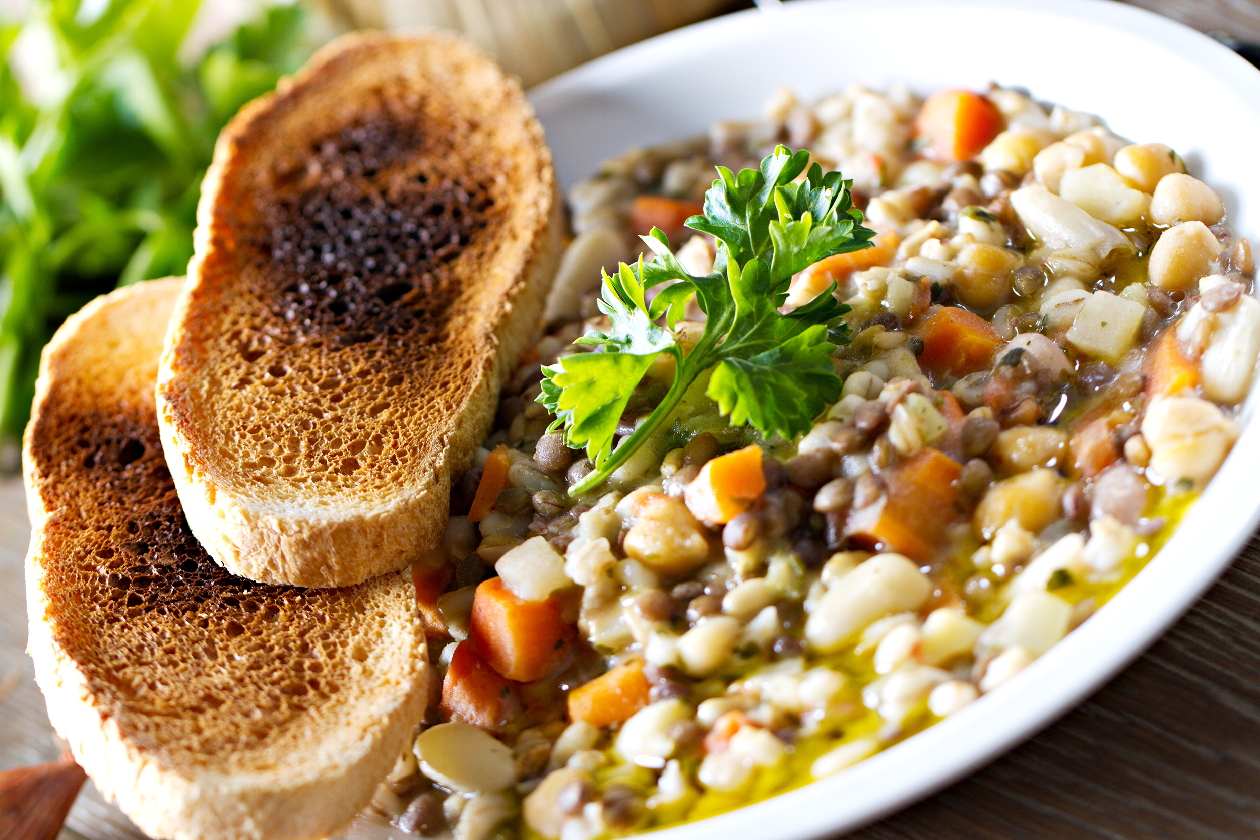 Chicken noodle soup in a white porcelain bowl, slight diagonal, with two pieces of french bread toast on the left, with a blurred background of parsley and a wooden table on the top left