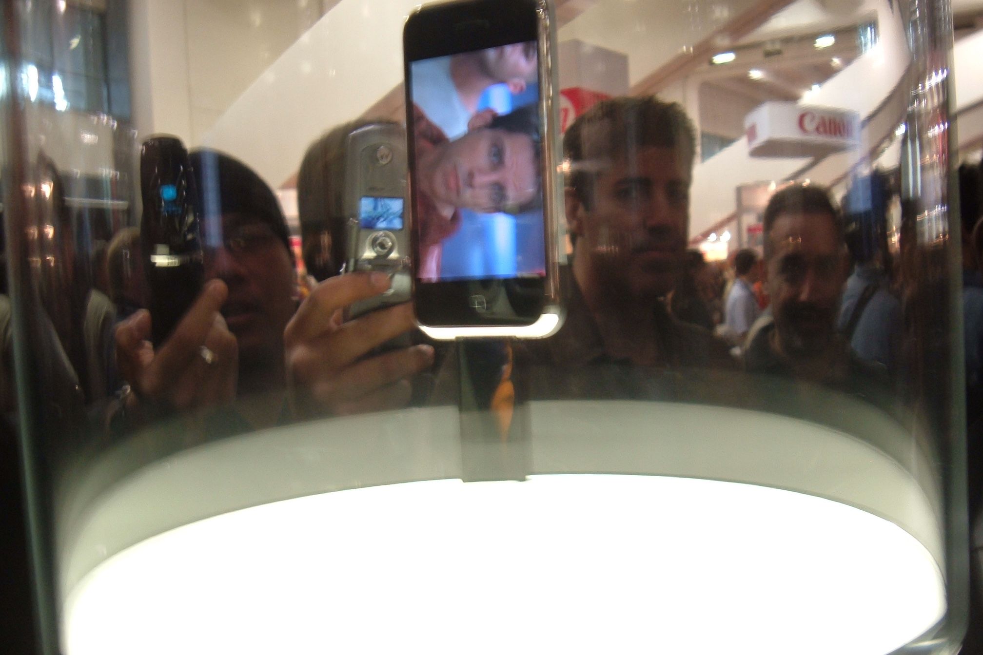Closeup of the first iPhone on display under glass and a brightly-lit light at the January 2007 Macworld Show, surrounded by several people looking at it and taking photos