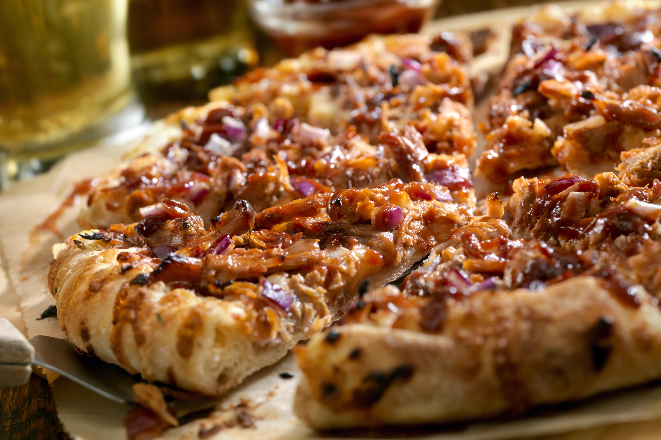 Closeup of BBQ pulled pork pizza, selective focus, cut into slices, one slice is on a pizza server, on brown wax paper, a glass of beer blurred in the background