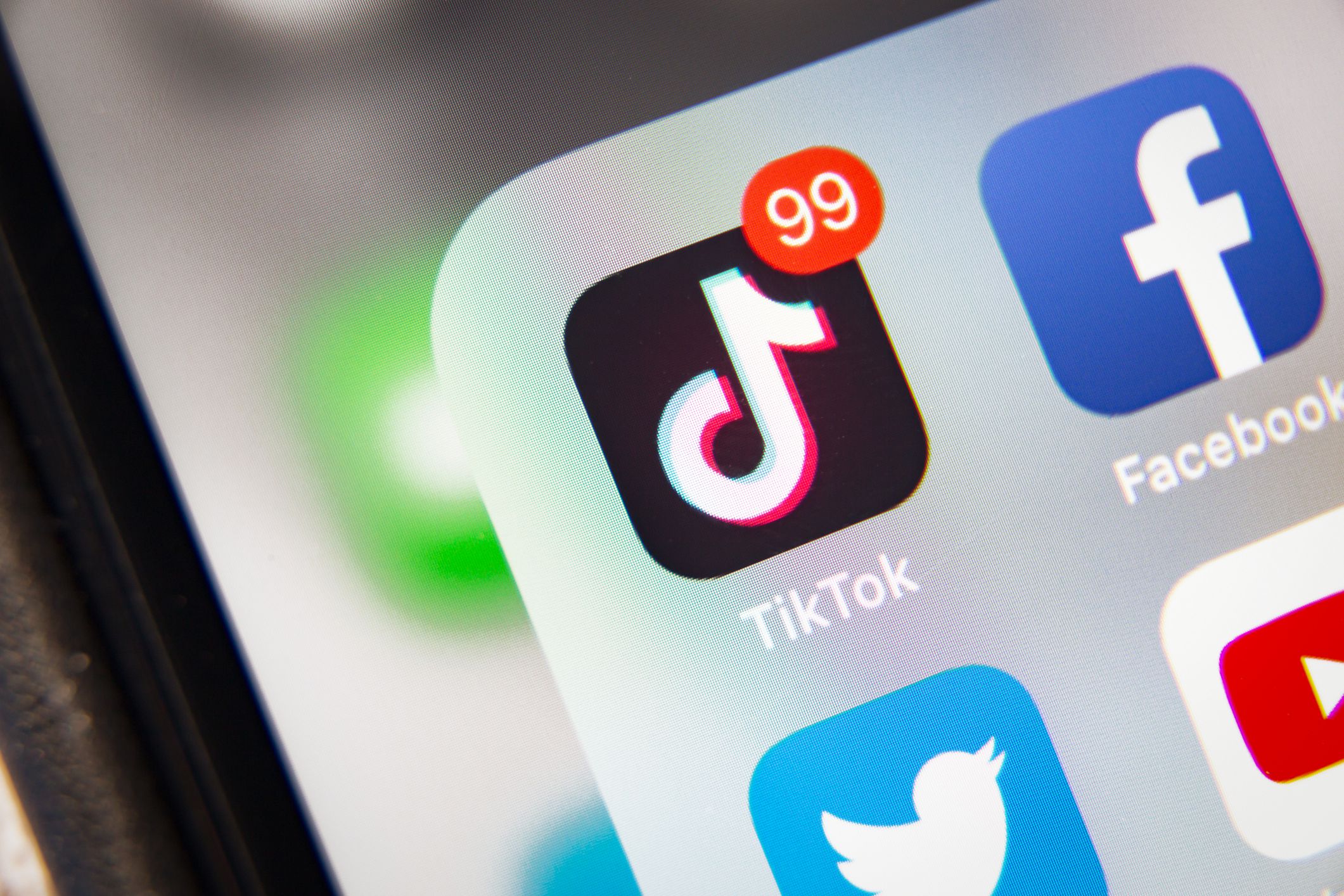 Closeup of TikTok app on the screen of an Apple iPhone XR, next to Facebook, Twitter, and YouTube apps in the pop-up window, against a blurred grey screen