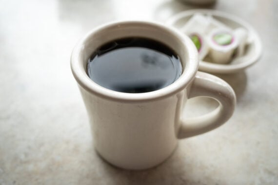 A white ceramic mug filled with black coffee—perfect for enjoying the best cheap coffee—sits on a light-colored surface, with small single-serve cream containers on a plate in the blurred background.