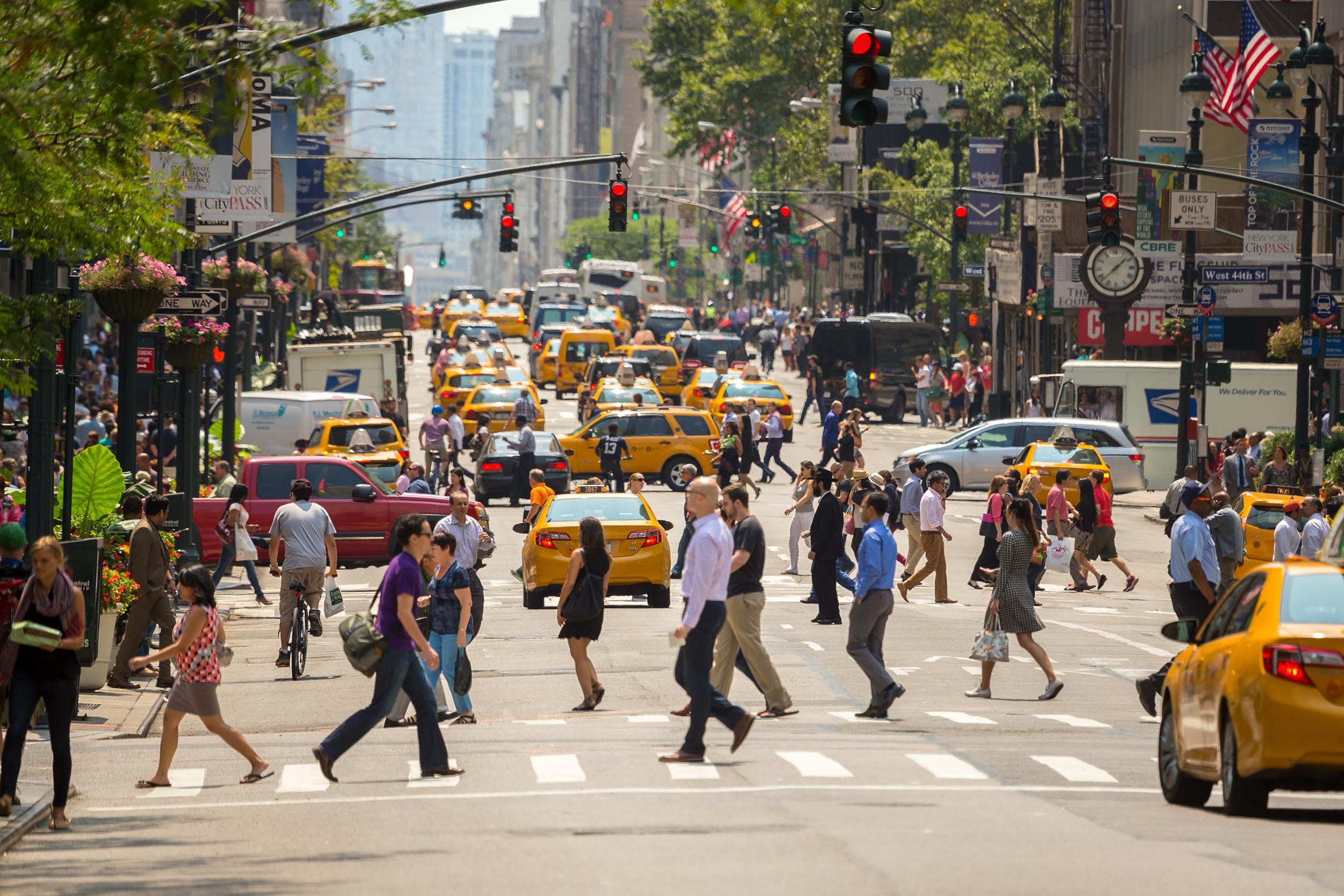 Crowd of people walking along a crosswalk in downtown Manhattan, street is filled with yellow cabs, trees and plants line both sides of the street, on a bright sunny day