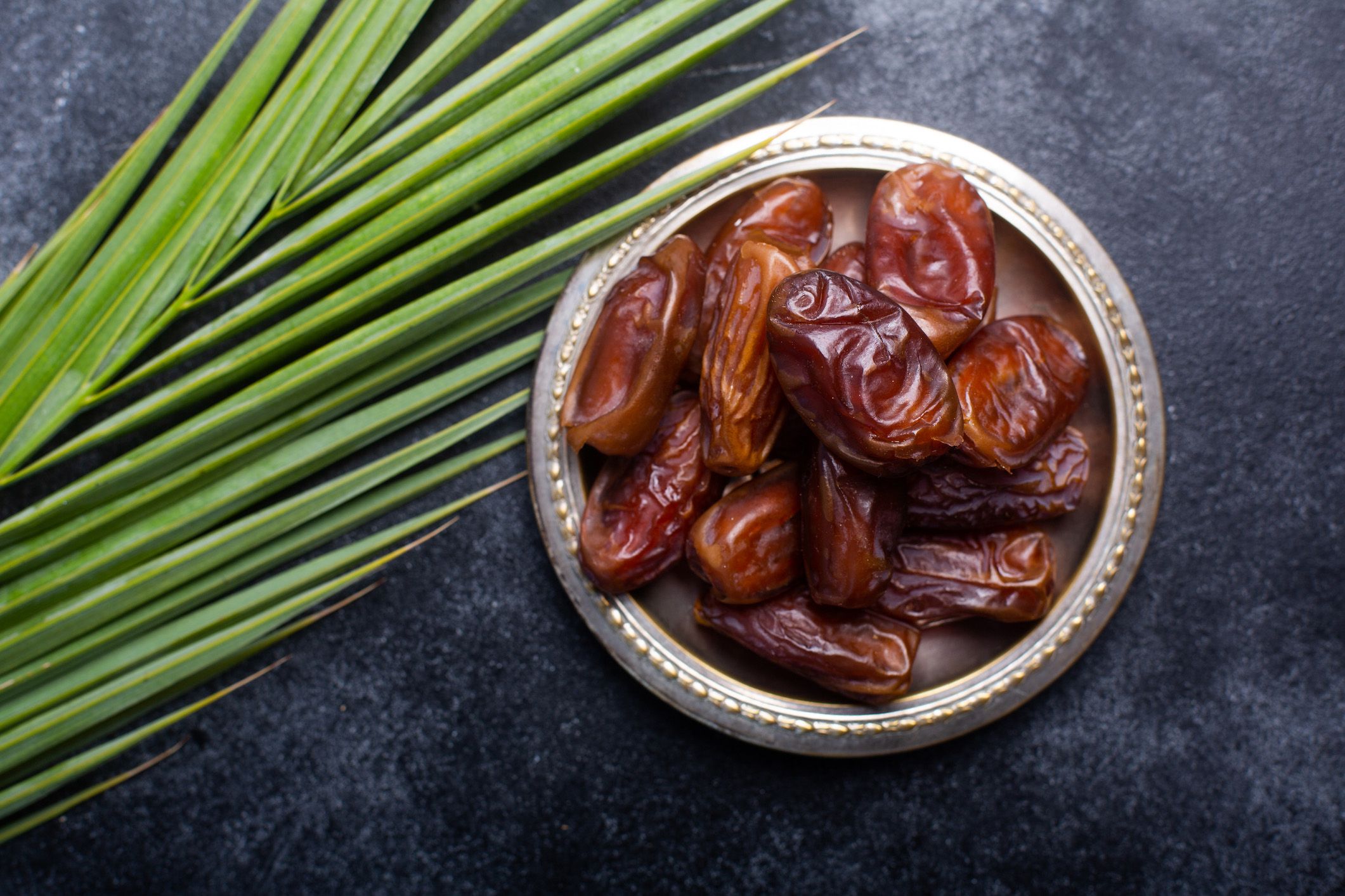 Dates in a silver serving bowl on a blue velvet background with a palm leaf