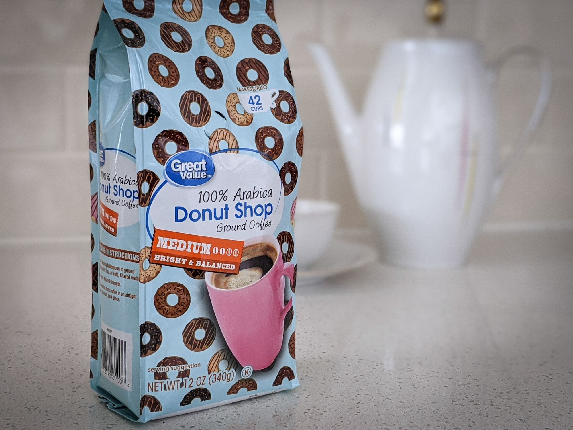 Great Value Donut Shop 100% Arabica Medium Roast to the left on a beige countertop, with a white porcelain kettle, tea cup, and saucer against a beige tile wall background