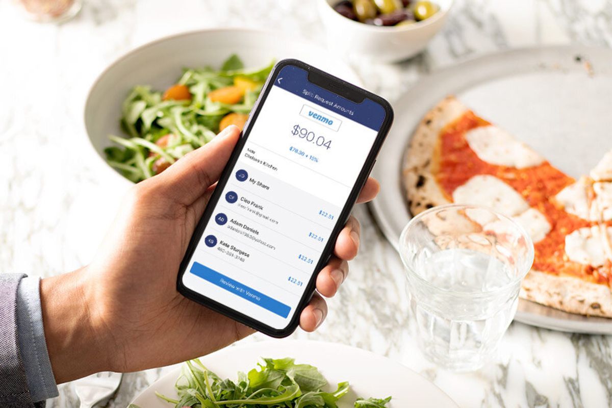 Left hand holding a smartphone with the Venmo app opened on the screen in the middle of the image, over a blurred array of several Italian lunch dishes over a white tablecloth