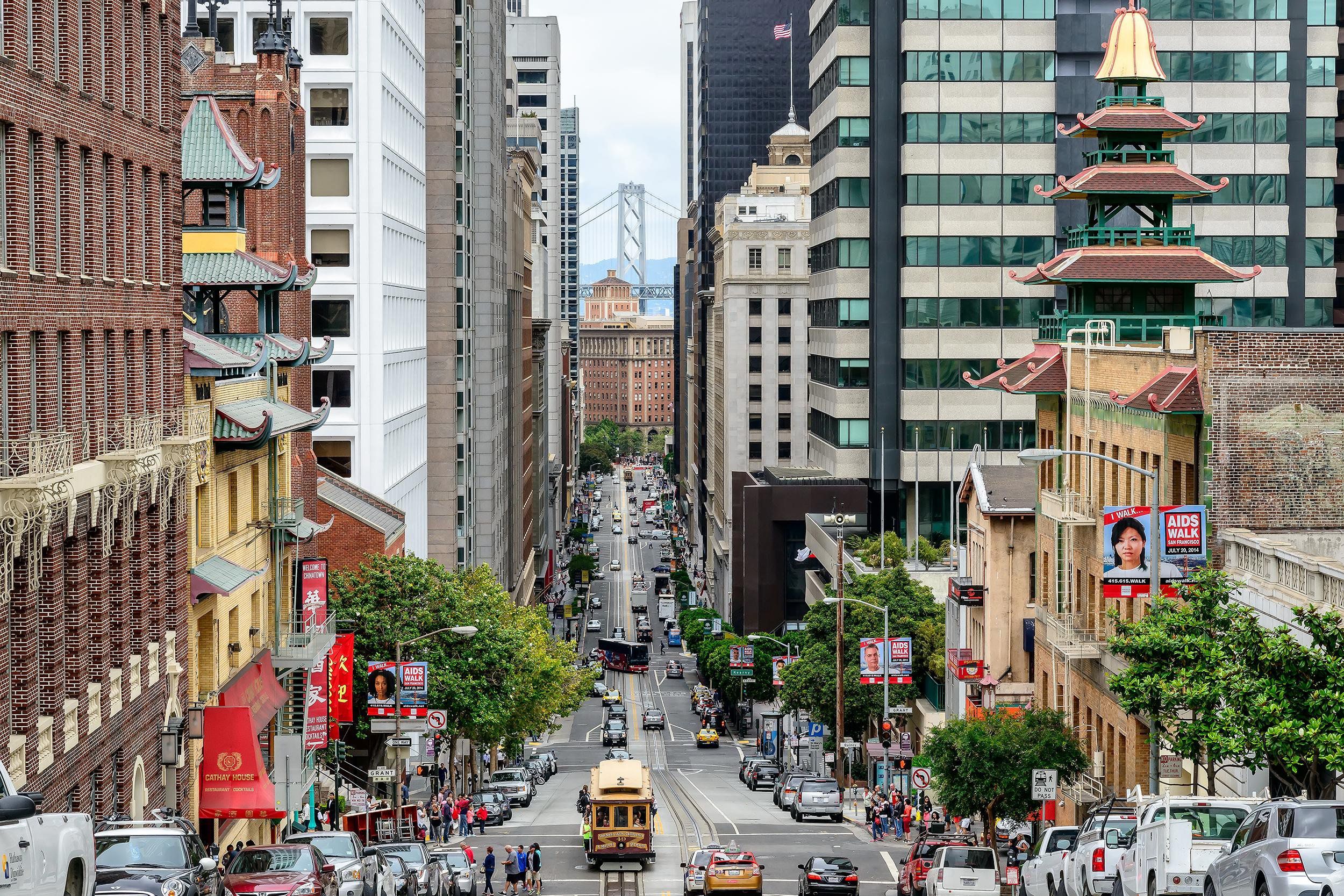 Hilly street in downtown San Francisco, cars line the road with very tall buildings, a cable car in the center, the Golden Gate Bridge in the distance, on a sunny day during summer