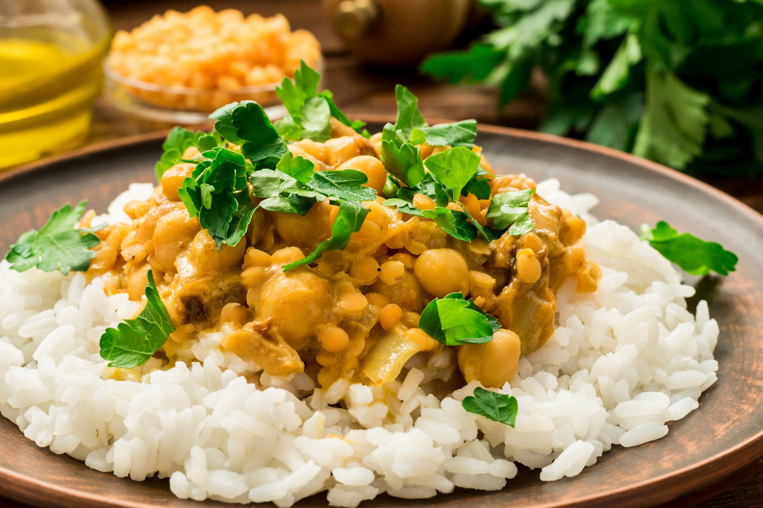Madras lentils with rice and veggies on a terracotta ceramic plate with a blurred background of a glass of oil, a glass bowl of lentils and more ingredients