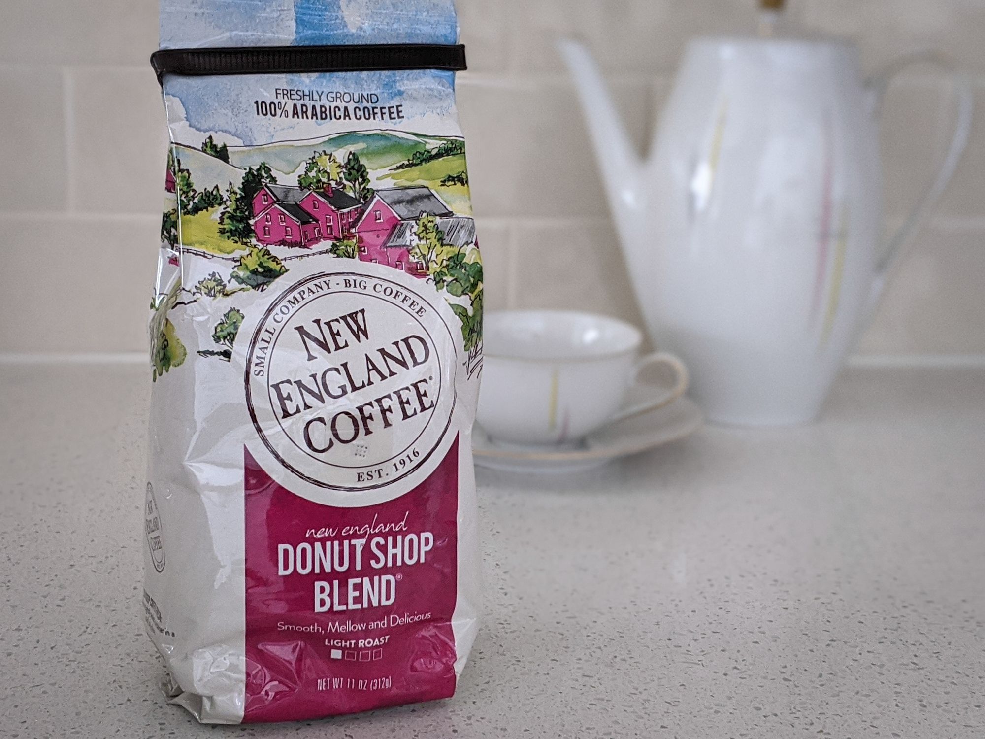 New England Coffee New England Donut Shop Blend to the left on a beige countertop, with a white porcelain kettle, tea cup, and saucer against a beige tile wall background