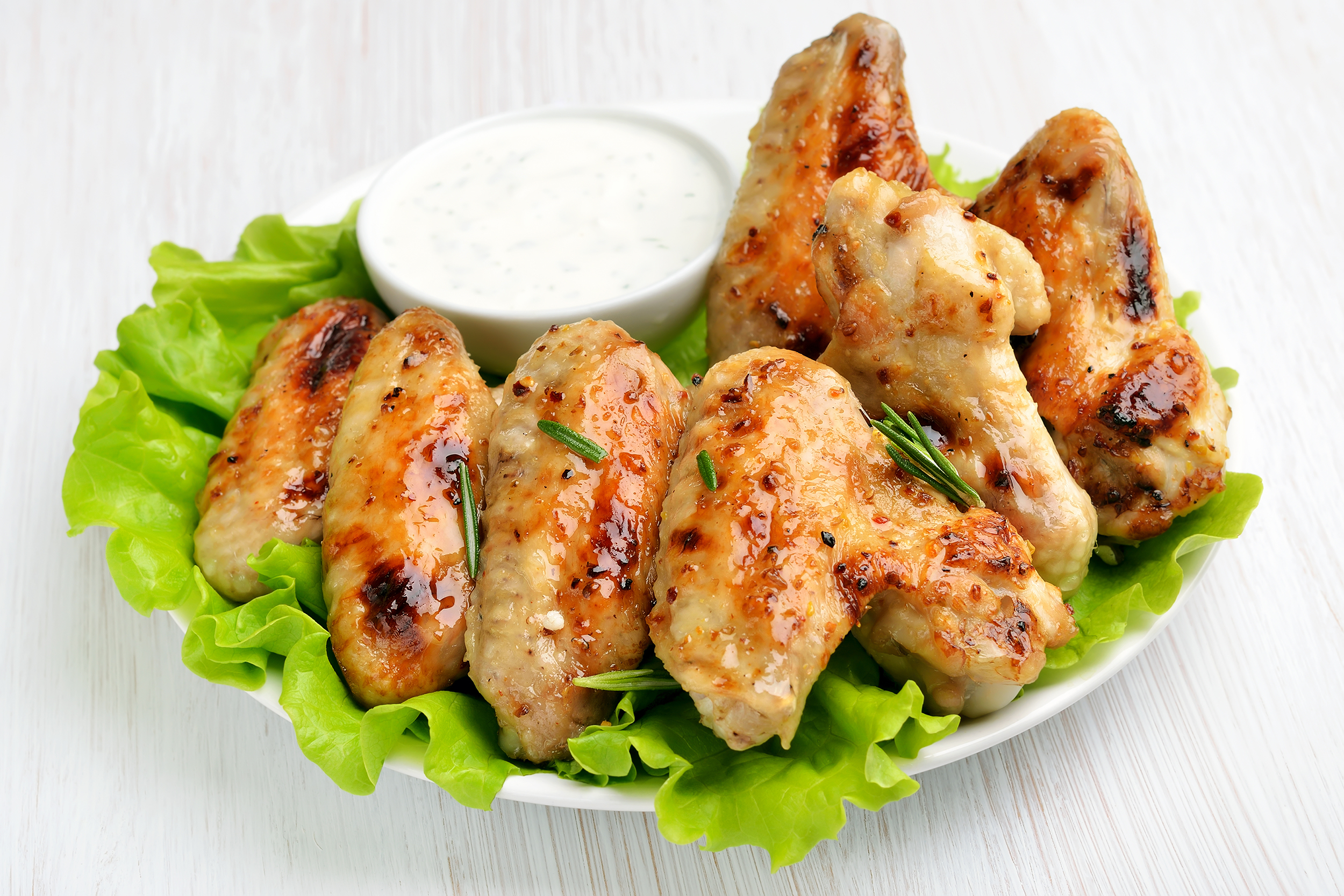 Parmesan and garlic baked chicken wings on a few lettuce leaves with a white dipping bowl of white sauce on a white porcelain plate on a white wooden table