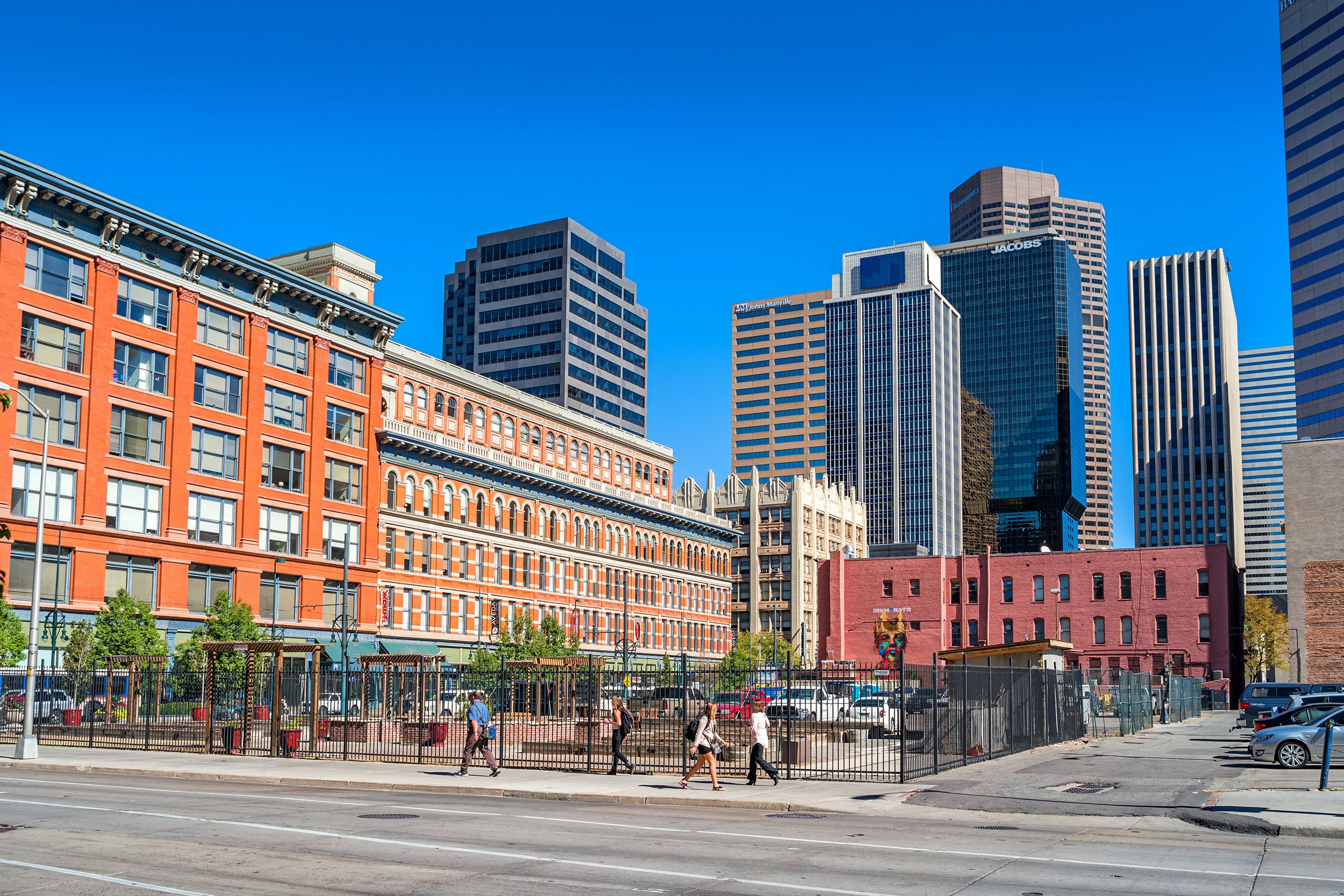 People walk along sidewalk of 15th Street in the Financial District in downtown Denver, road in the foreground, historic buildings and a parking lot, tall modern buildings in the background, on a bright sunny day in summer