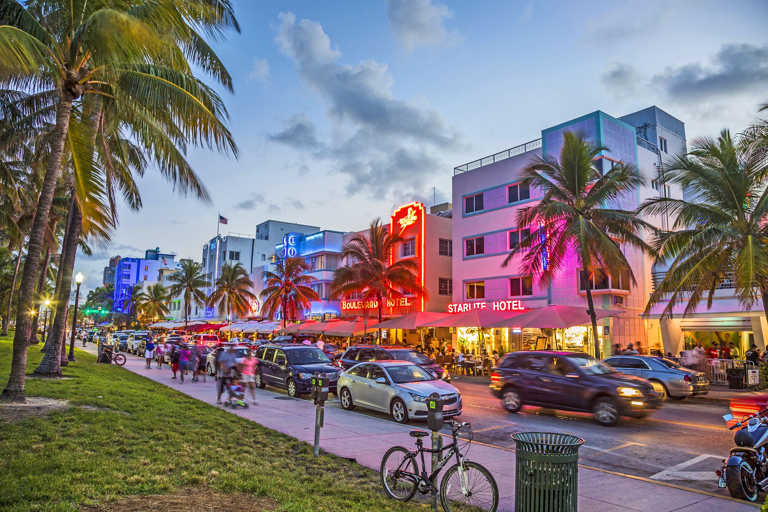 People walk along palm tree-lined sidewalk across the street of art deco buildings on Ocean Drive, Miami during sunset, colorful lights on the buildings