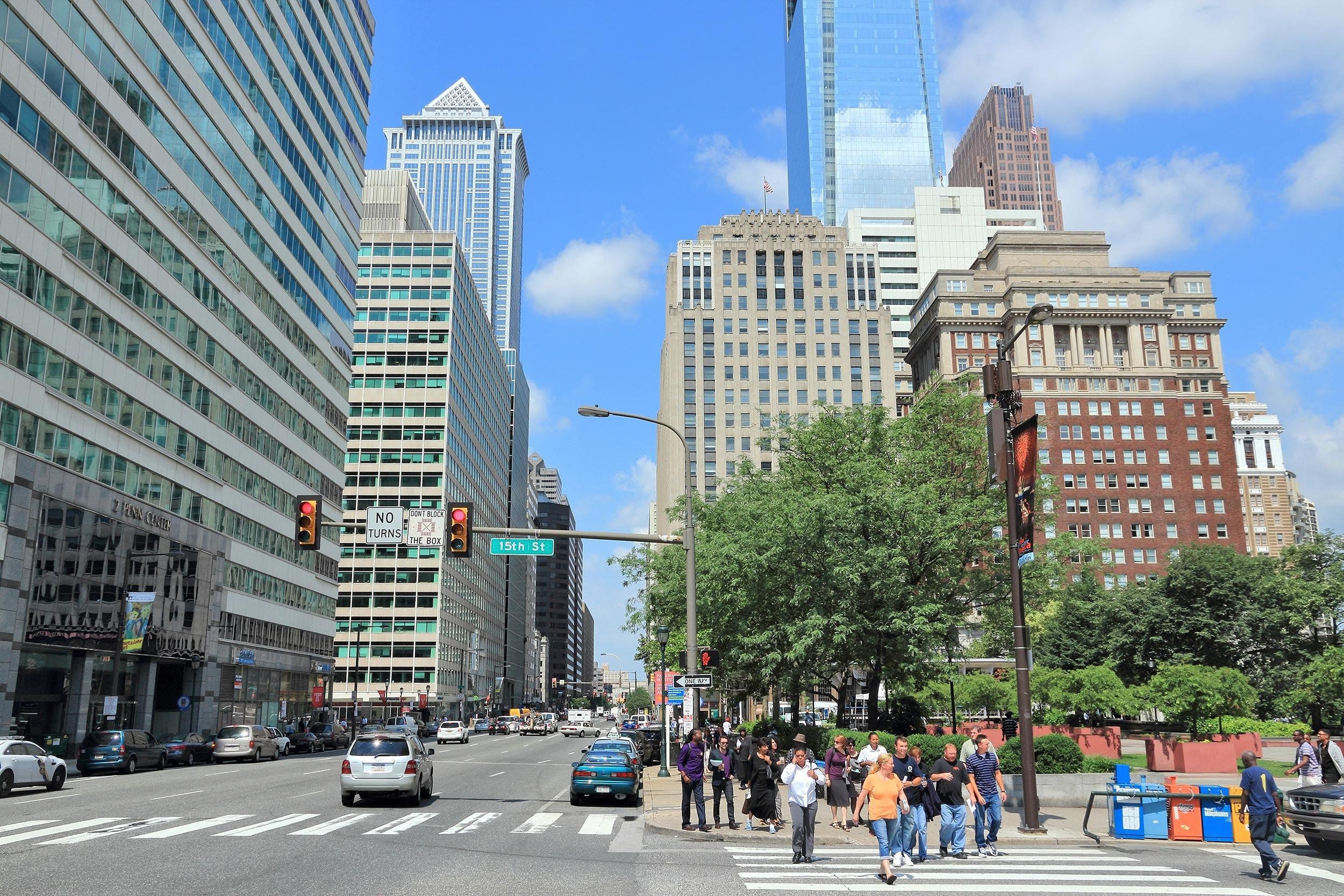 People walking along crosswalk in downtown Philadelphia, road on the left, crosswalk on the right with park behind it, tall buildings line the road, on a bright sunny day in summer
