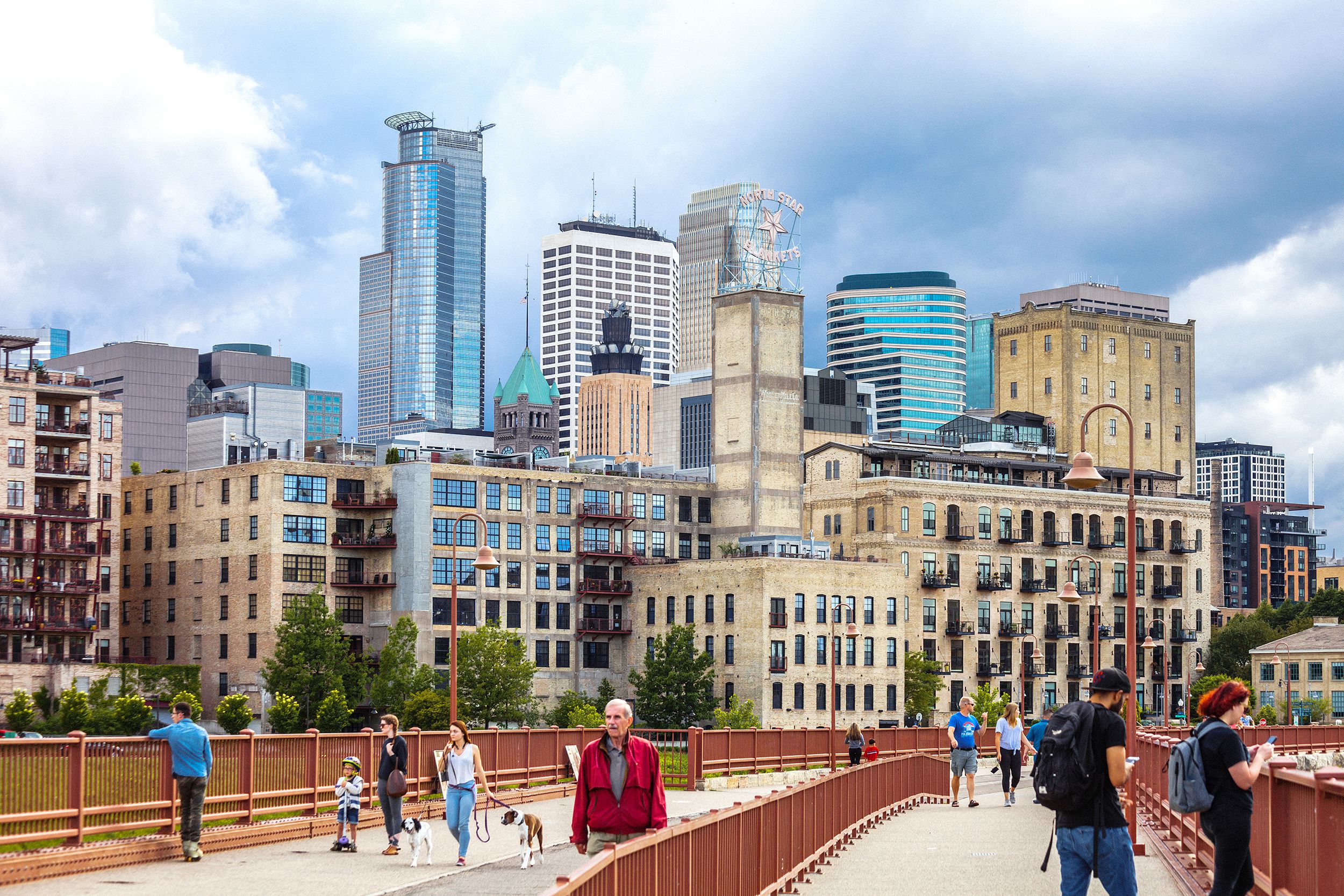 People walking on the Stone Arch Bridge to cross the Mississippi River in downtown Minneapolis, in the foreground, high rise buildings in the background, on a summer day