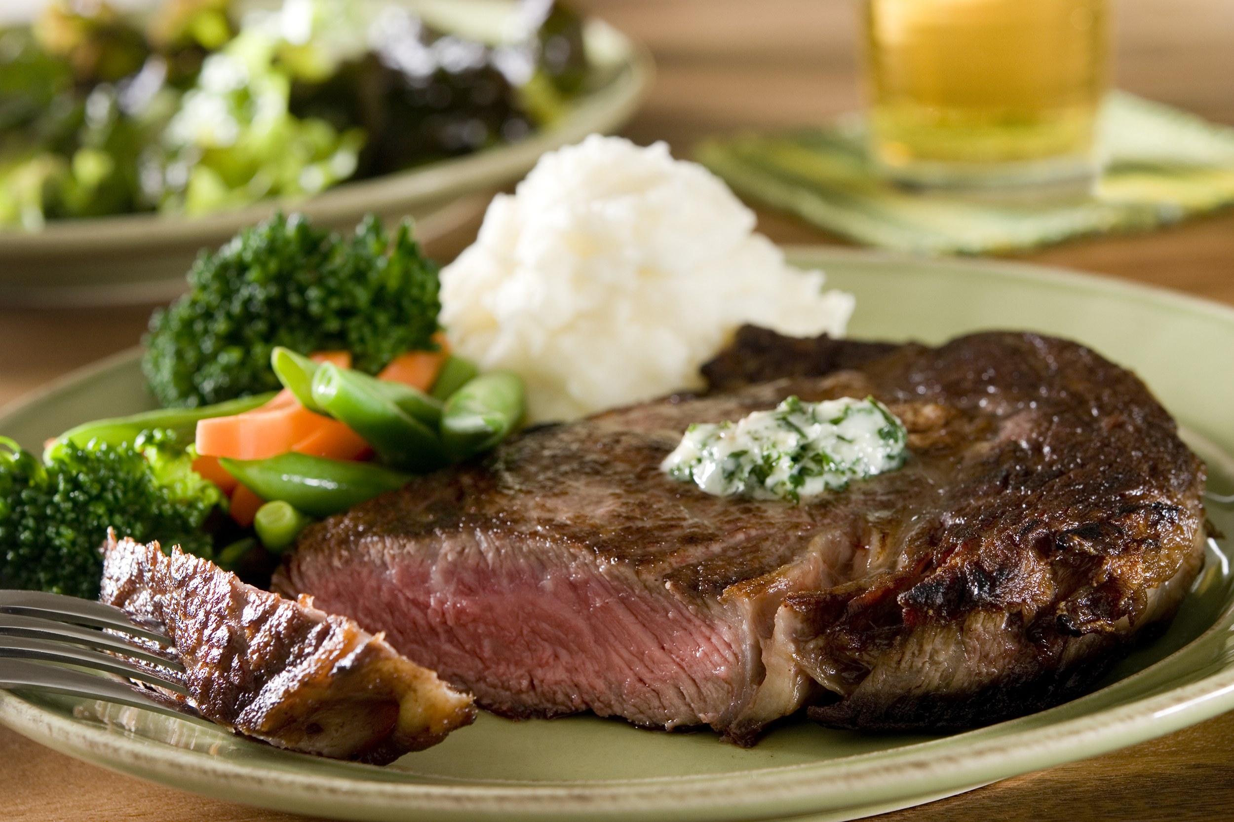 Prime rib-eye steak with mashed potatoes on a beige ceramic plate, one piece of steak on a fork placed on the left, with a blurred background of a salad on a beige ceramic plate and a glass of beer