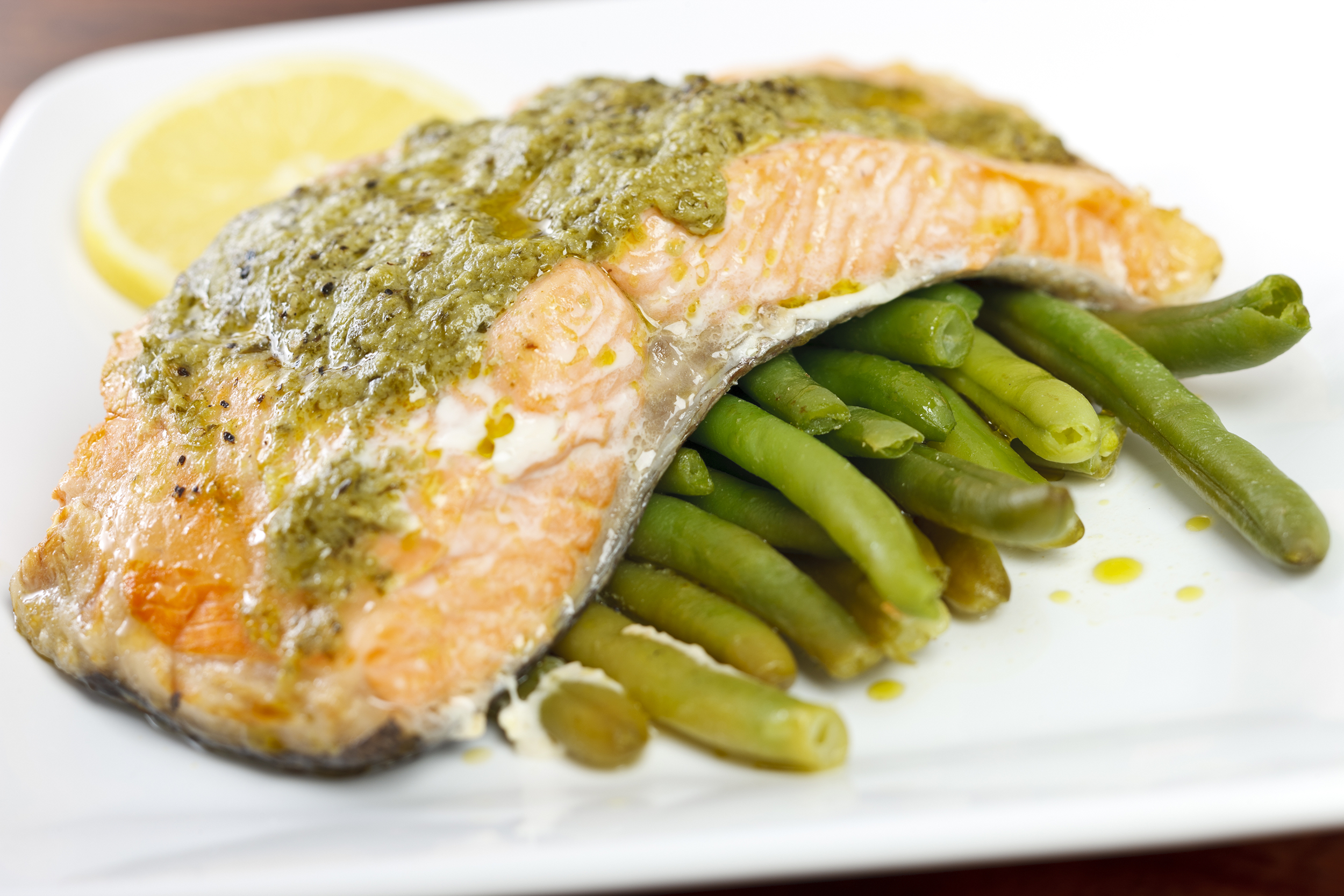 Salmon Milano with basil pesto butter over green beans on a square white porcelain plate, selective focus, a slice of lemon on the top of the plate, blurred background of a dark brown table along the corners