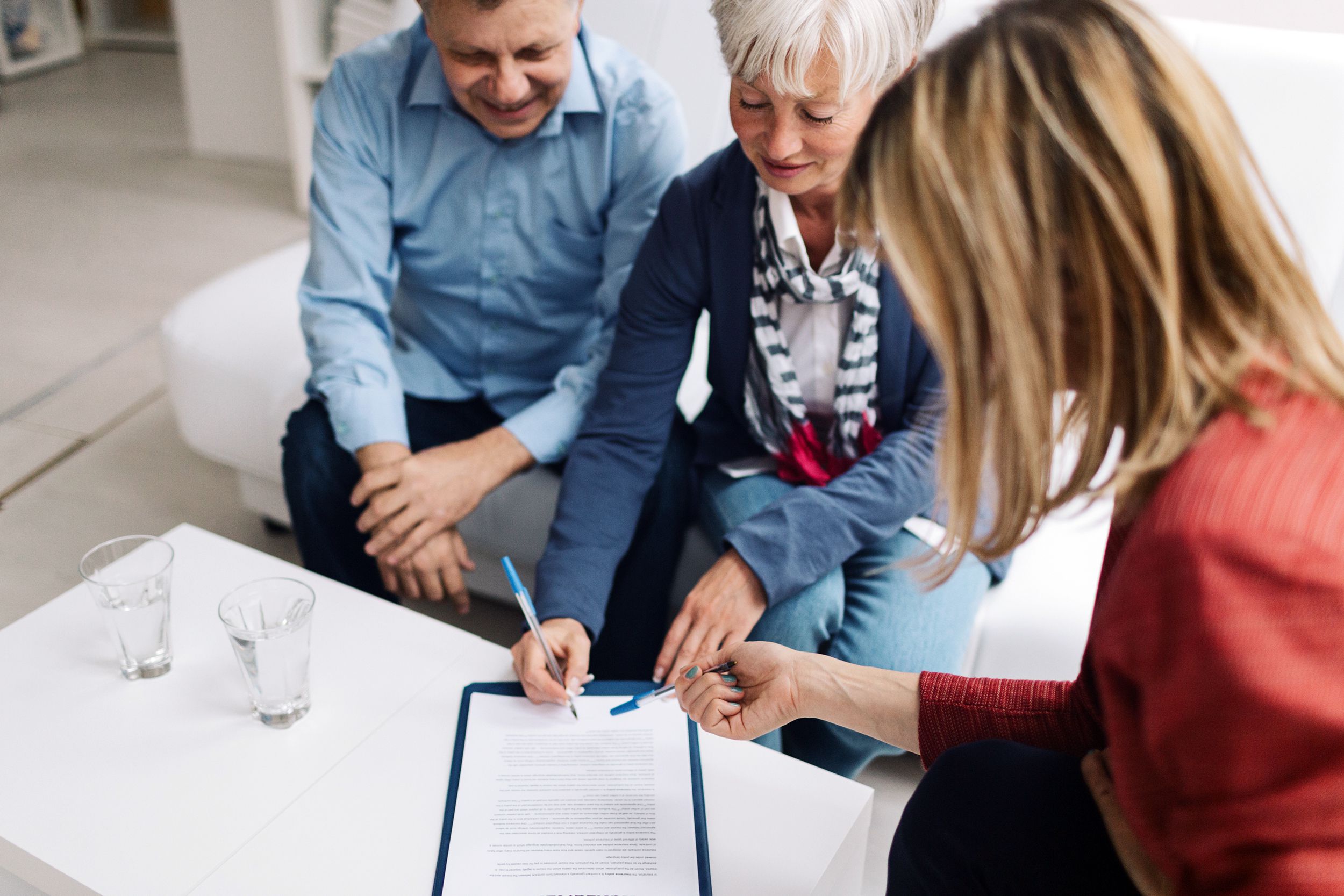 Senior couple having a meeting with a female financial advisor, senior woman is holding pen and signing contract while senior man looks on, financial advisor is pointing to contract with her pen
