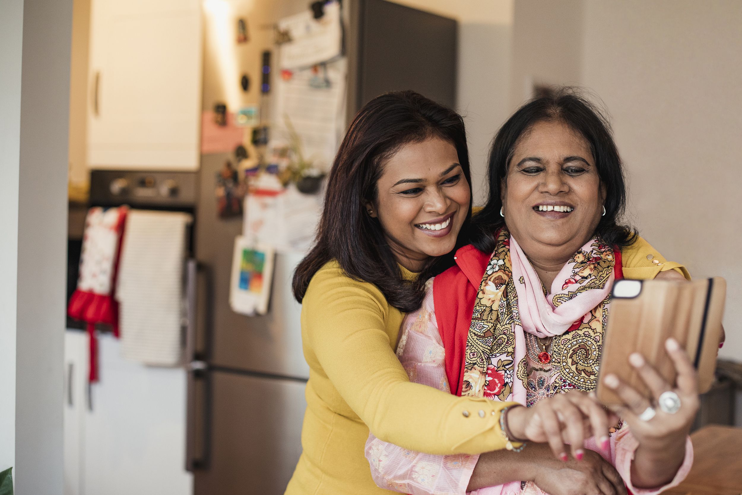 Senior mother and daughter taking selfie in living room, mother is holding smartphone, kitchen appliances are in the background