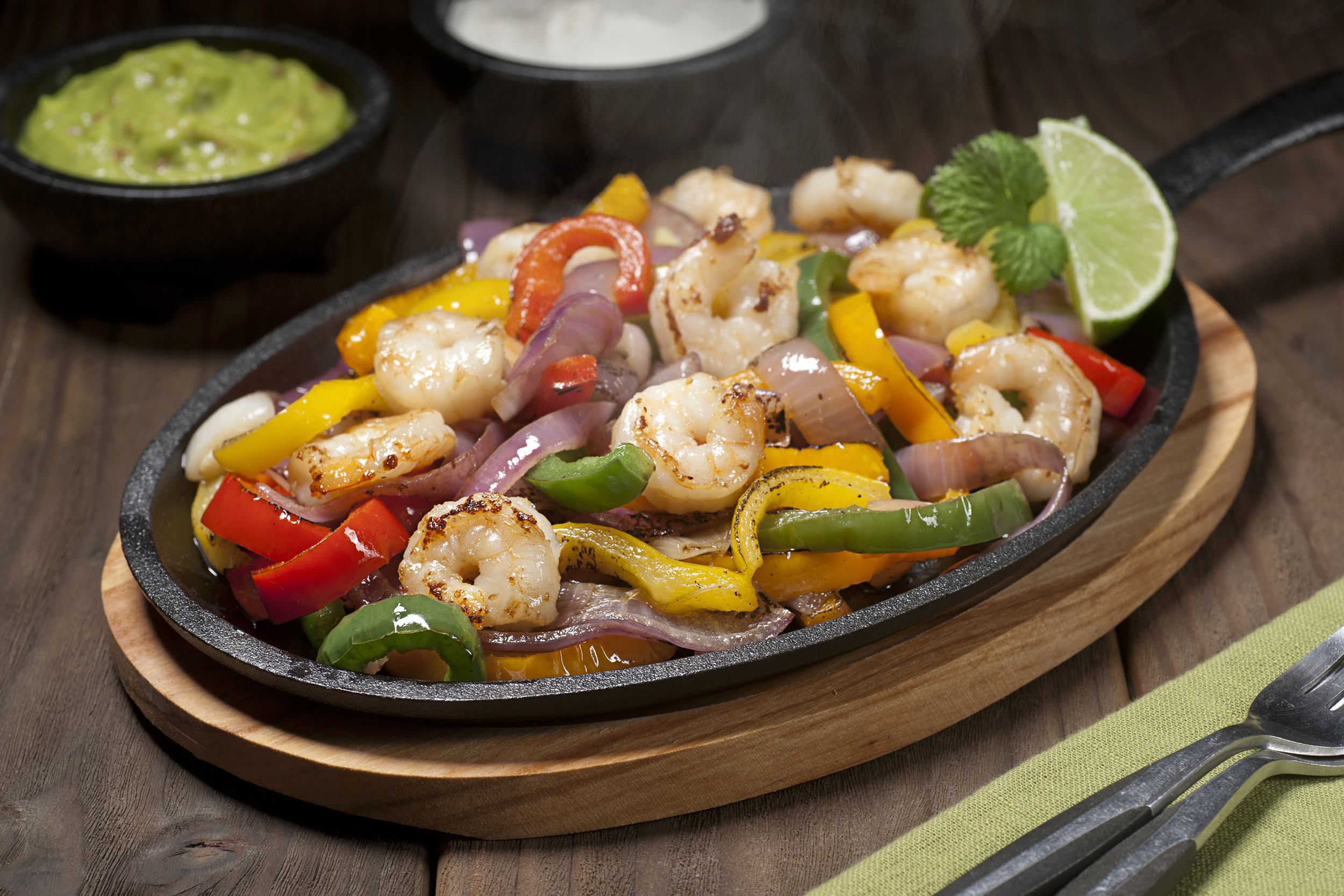 Shrimp fajitas in a cast iron skillet on a wooden serving board on a wooden table with an avocado green napkin and utensils on the right, two dipping cups of guacamole and a white sauce, with a blurred background