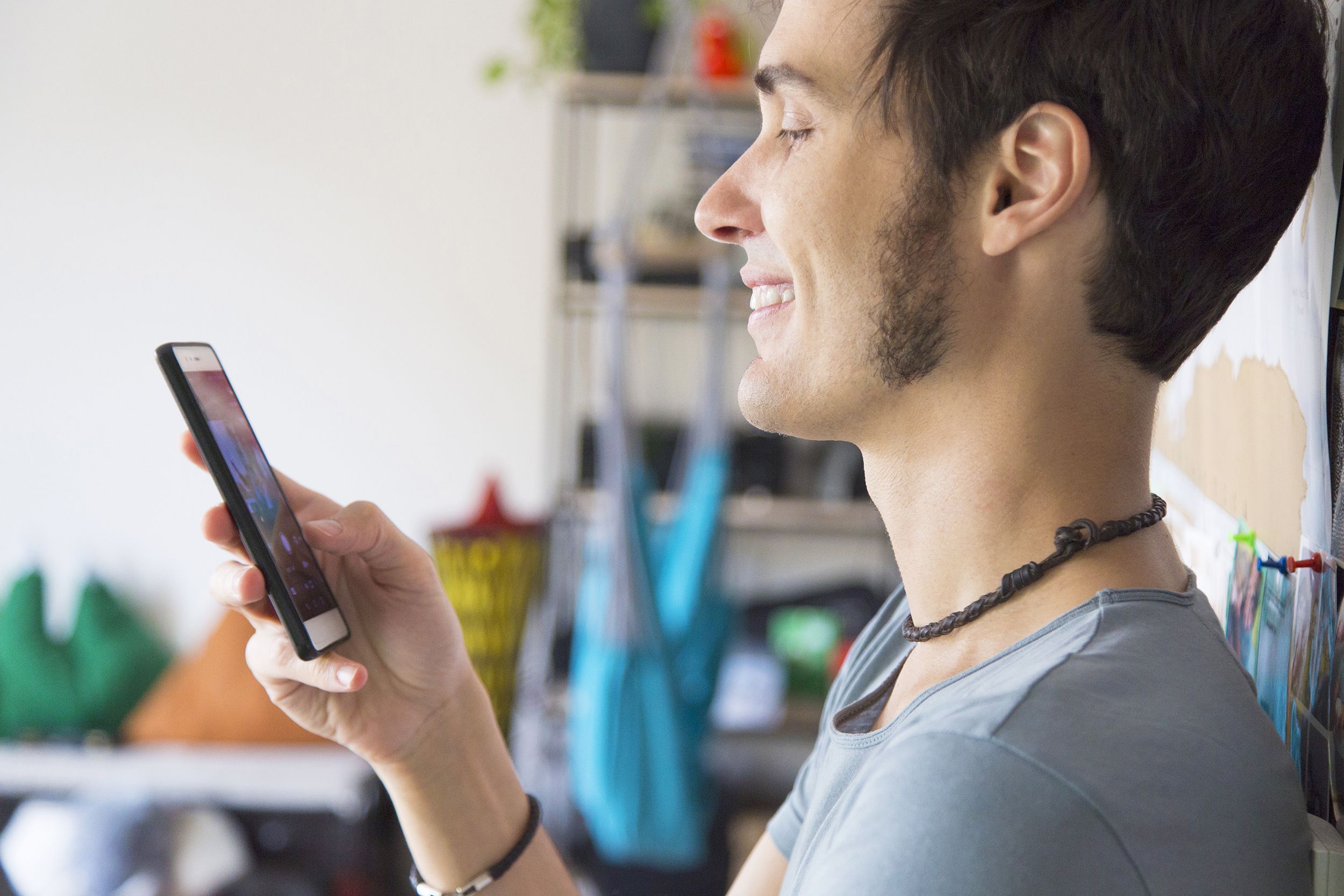 Sideview of the top of a young man smiling as he is using his smartphone, holding it in his right hand, standing against a large drawing, with the rest of his art studio blurred in the background