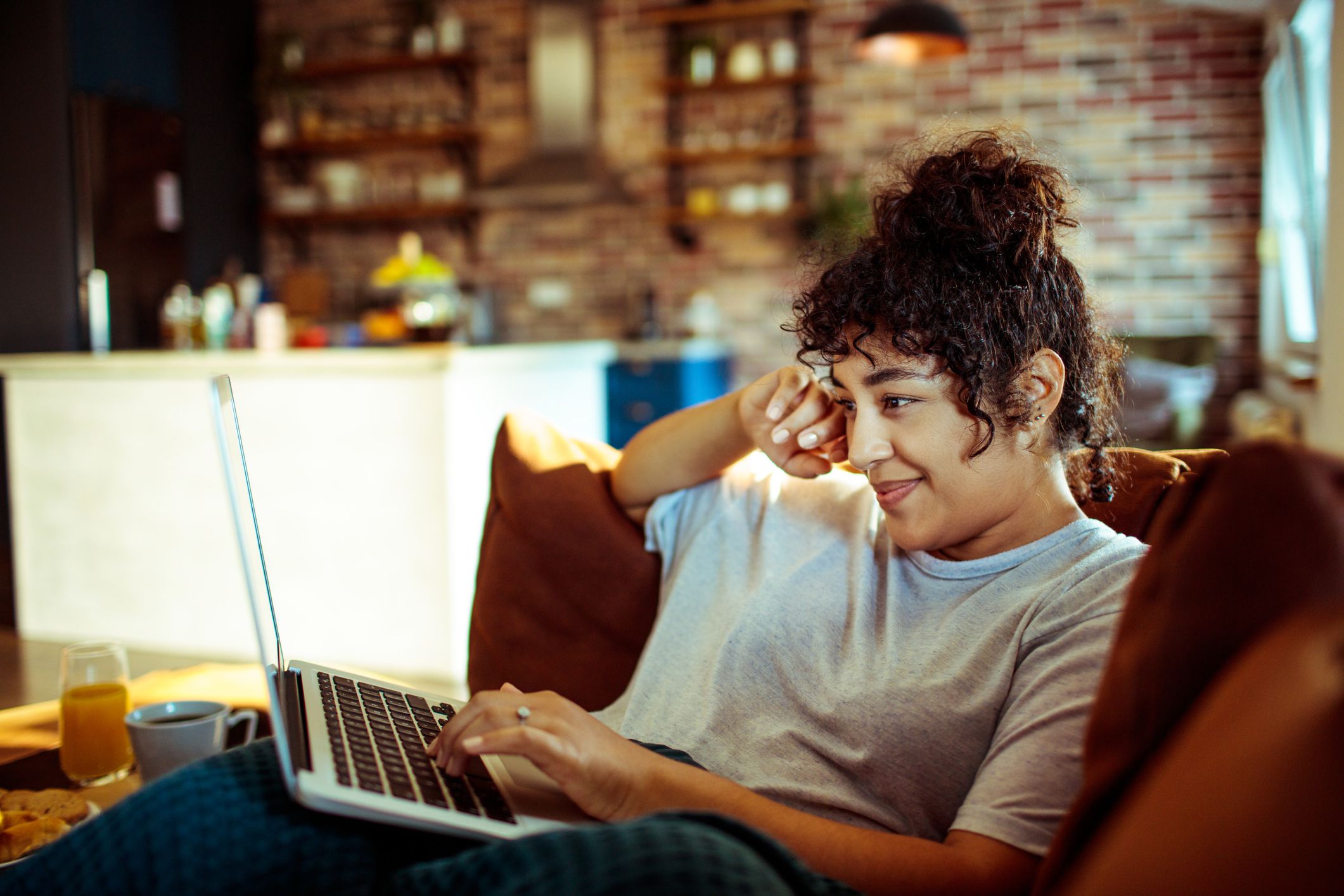 Sideview of a young woman laying on a couch with a laptop on her lap, she is looking at the laptop, with a blurred background of the living room and kitchen of a small city apartment