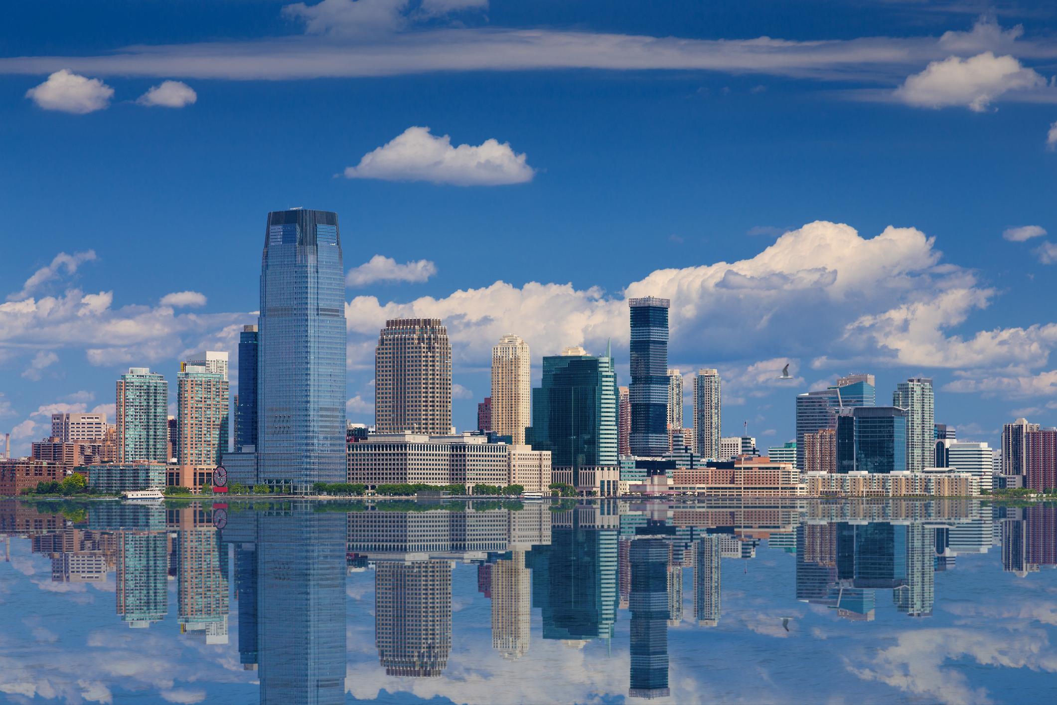 Skyline of Jersey City, New Jersey, with Goldman Sachs Tower, blue sky, and clouds reflected reflected in water of Hudson River
