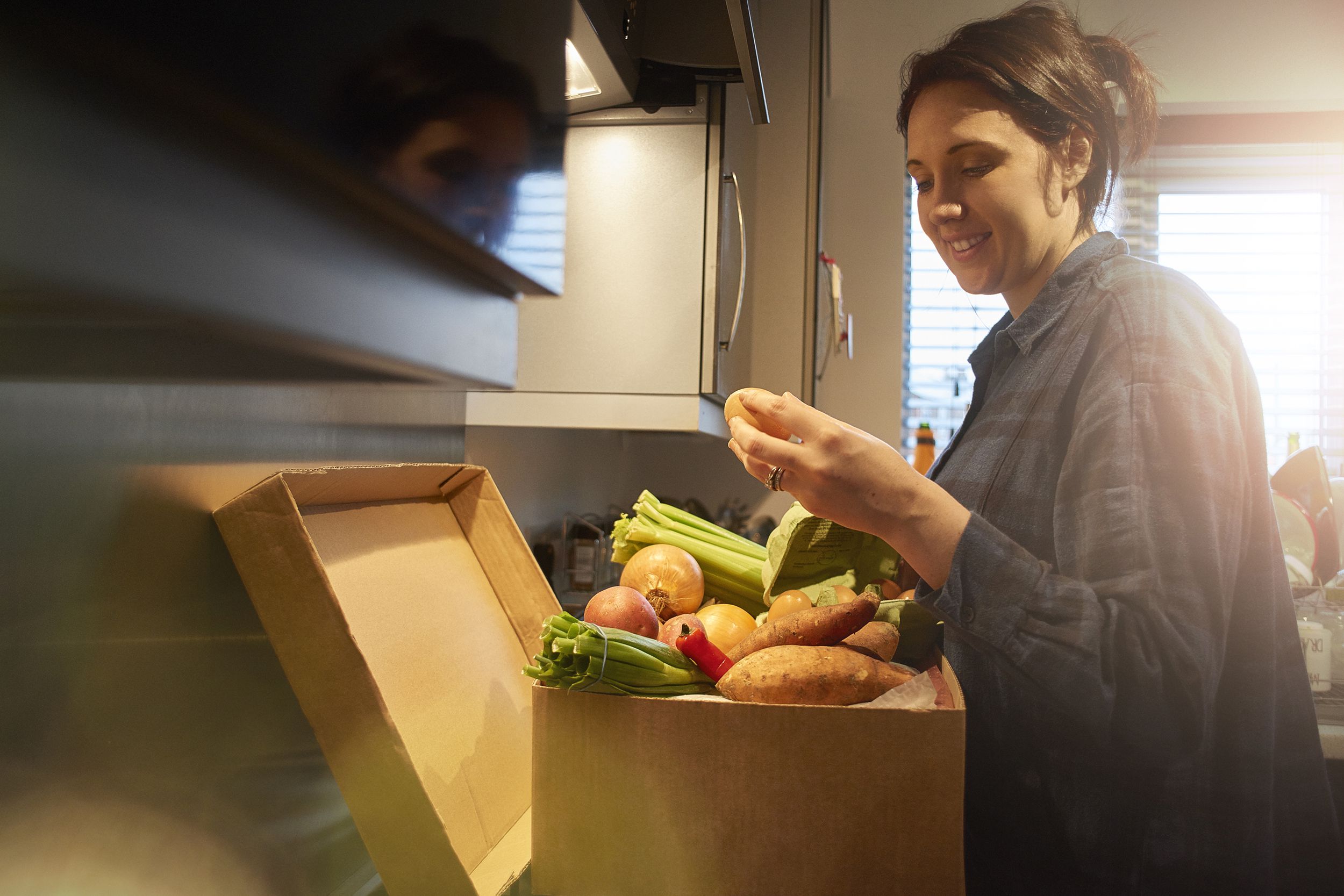 Smiling young woman checks over eggs and vegetables in a box from a delivery, in her kitchen, box is laying on the counter with the sink and windows in the background