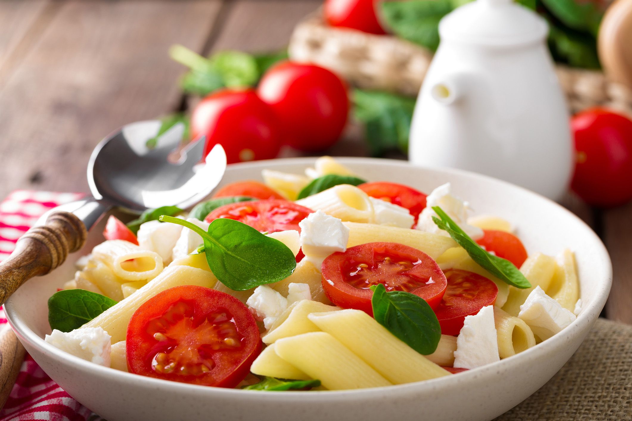 Spinach tomato feta pasta in a white bowl with a steel and wood serving spoon