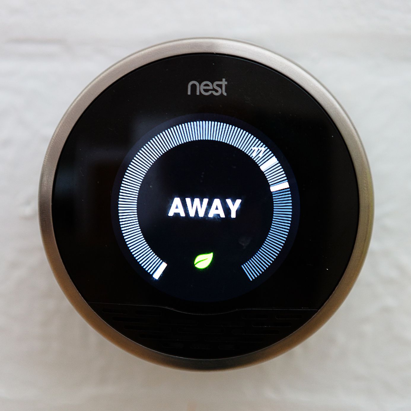 The Nest Learning Thermostat in away mode, chrome colored, on a white wall blurred