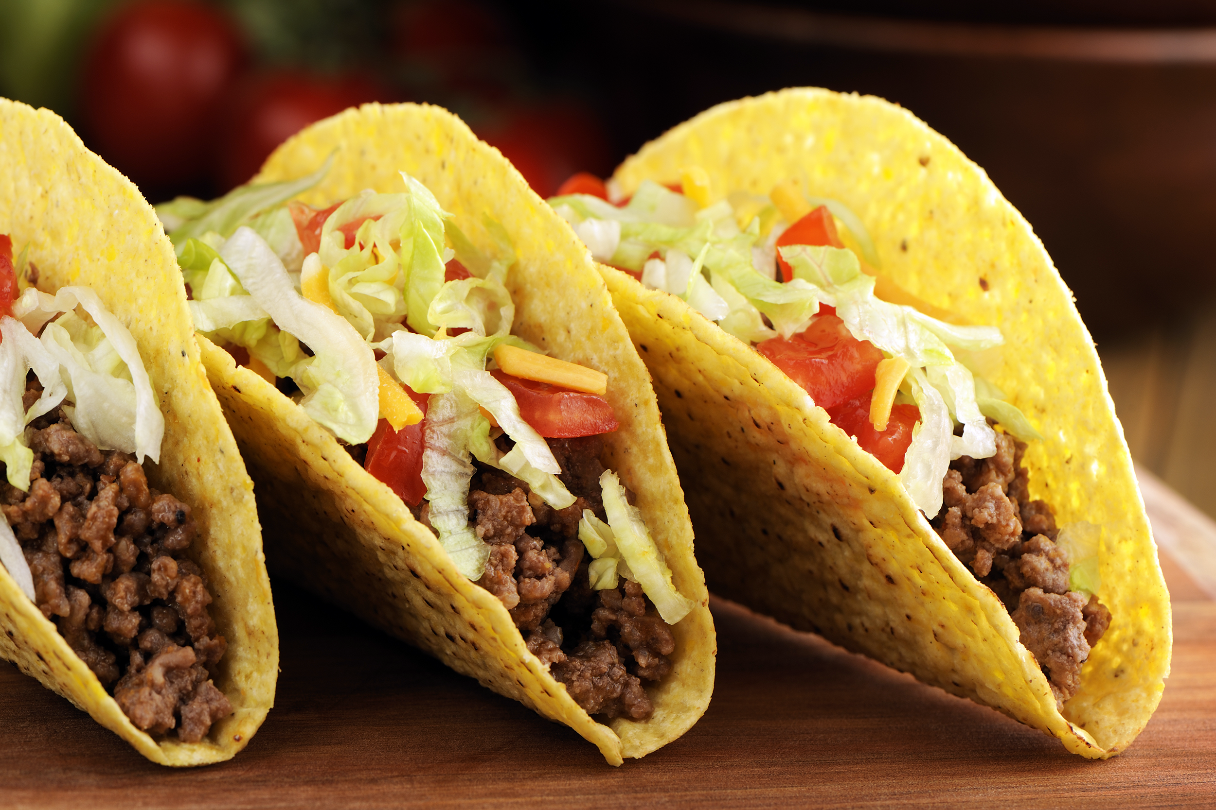 Three standing Tex-Mex organic ground beef tacos slightly leaning on each other, on a bamboo cutting board with a blurred background of dark browns