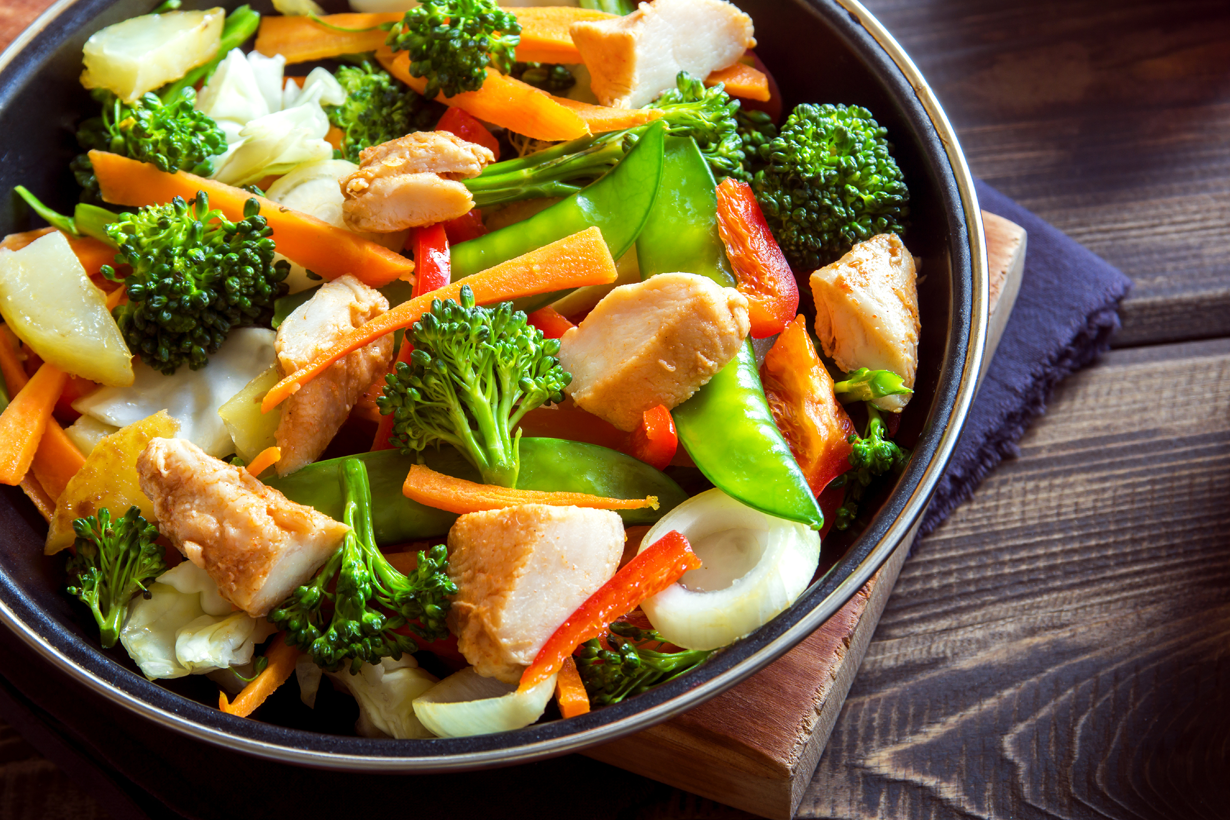 Top-view of chicken stir fry in a dark brown ceramic bowl, on the left, on a wooden board and a navy napkin on a wooden table, on the right