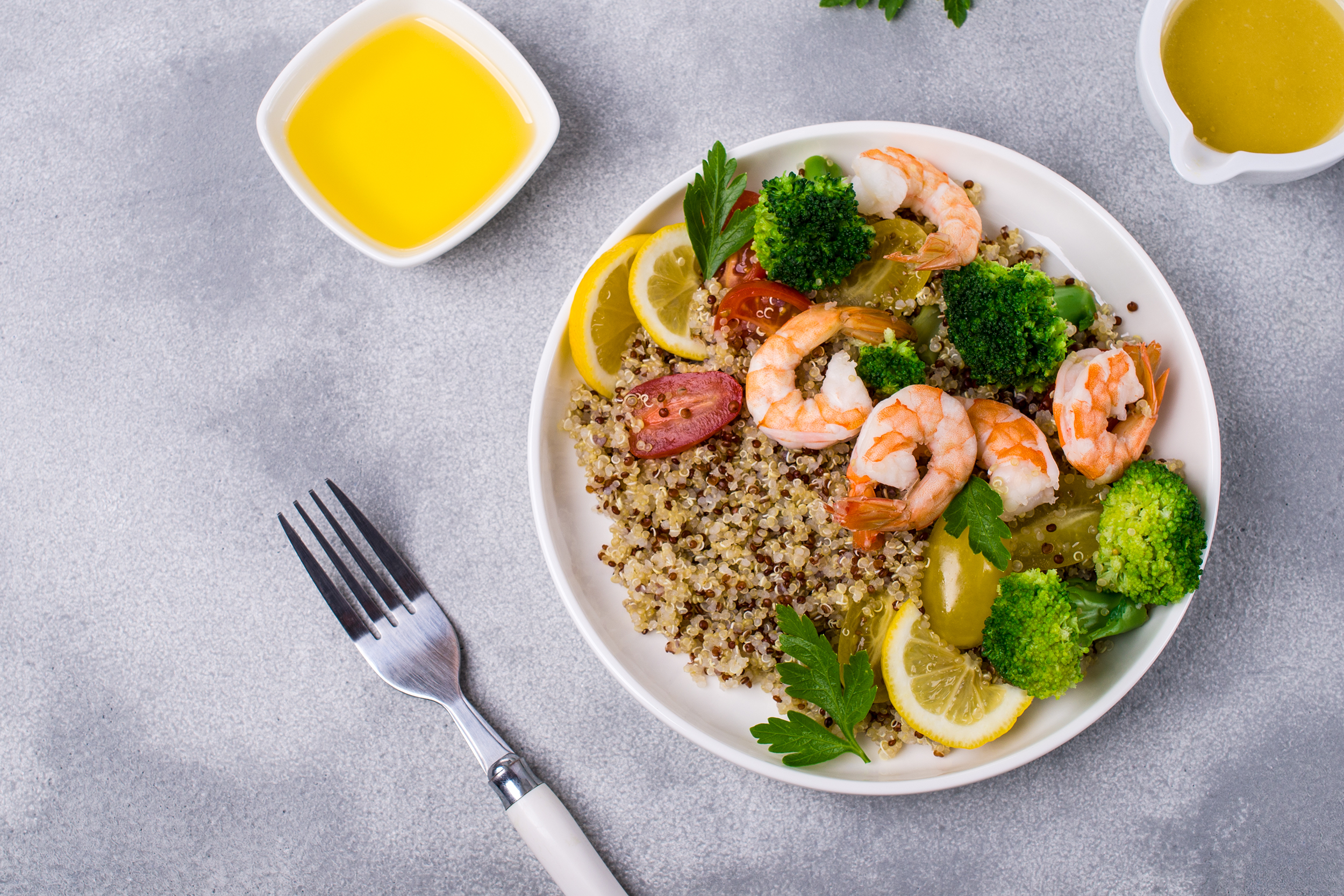 Top-view of shrimp and quinoa on a white porcelain plate on the right, with a fork on the bottom middle and a small white dipping bowl of butter on the top middle, on a grey table