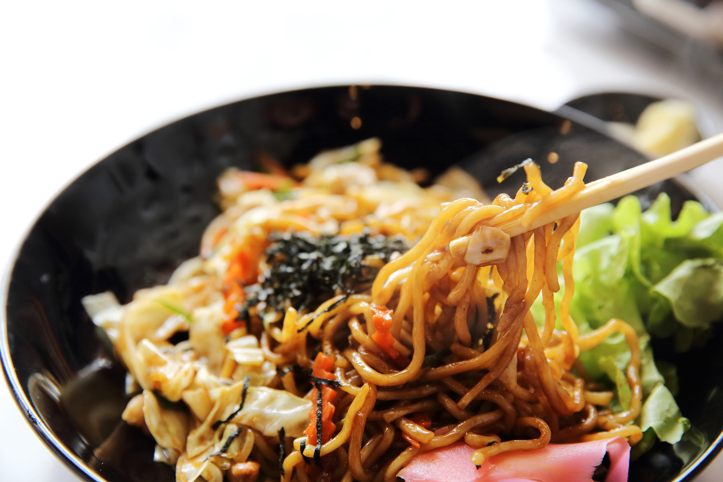 Tuna yakisoba with vegetables with chopsticks picking up some, selective focus, in a black porcelain bowl on a white background