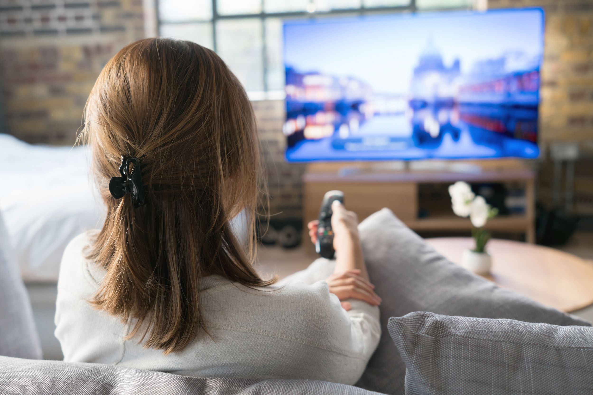 Back of woman sitting on couch with remote pointed towards an ultra high-definition TV across the living room of her home, selective focus, brick walls and window showcasing cityscape