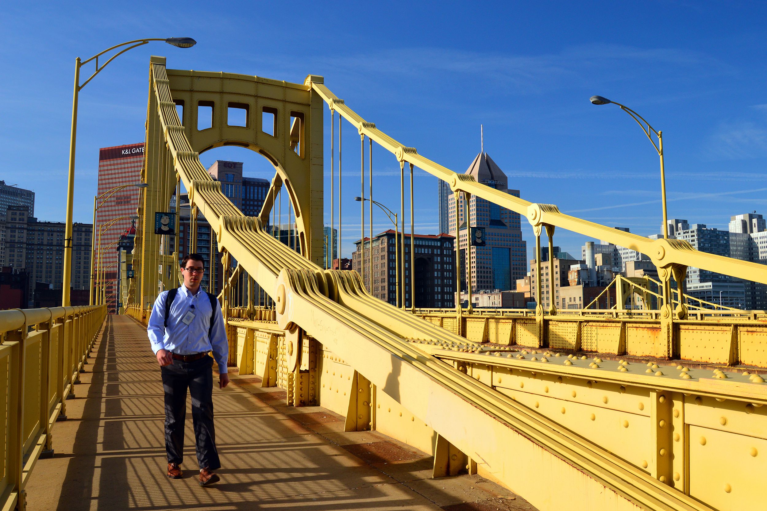 A young man walks across the Roberto Clemente Bridge in Pittsburgh, during his morning commute, light yellow bridge with skyline in the background, a blue sky