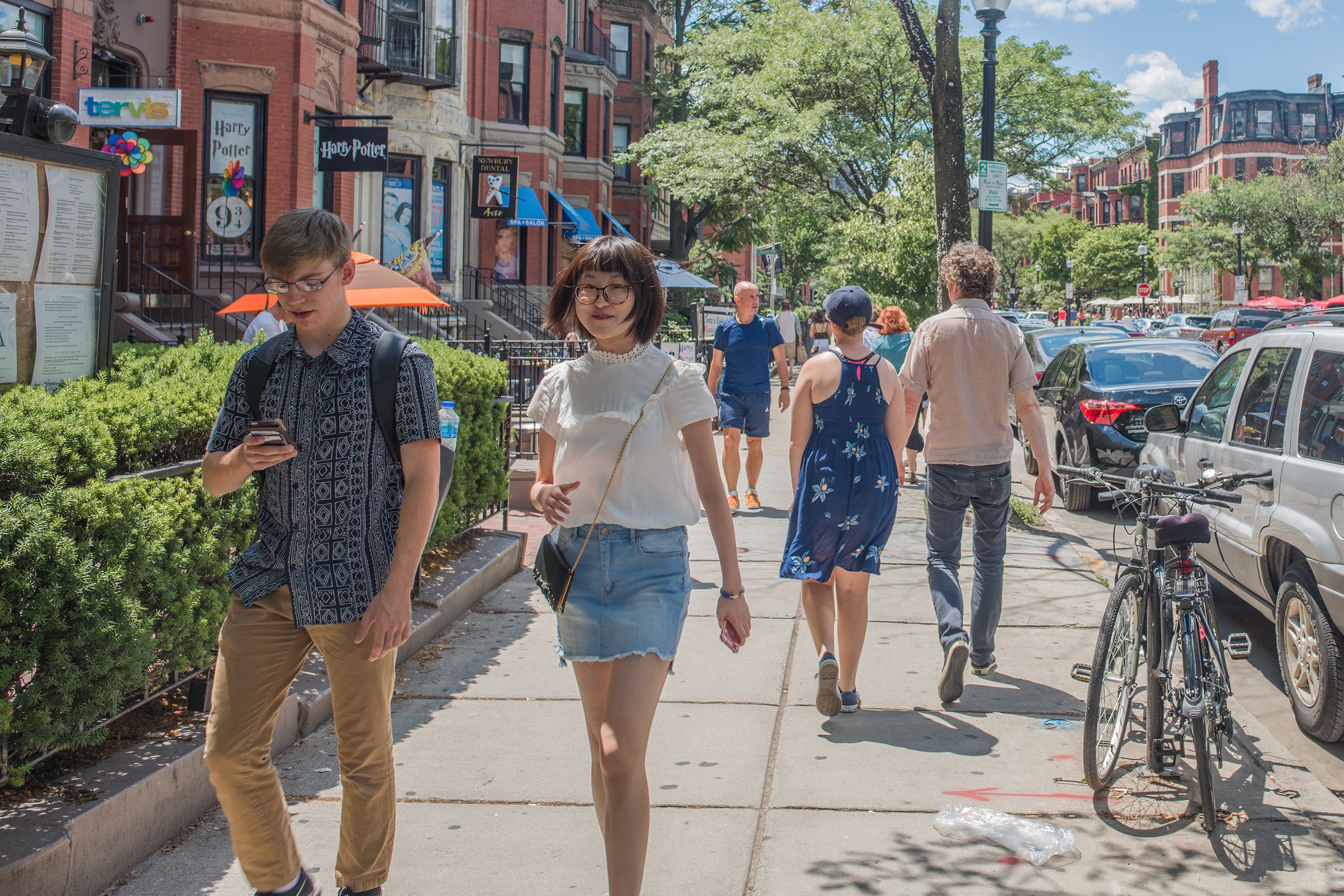 Young people walking along sidewalk in Back Bay neighborhood, Boston, one woman is looking at the camera, historic buildings and trees, on a bright summer day