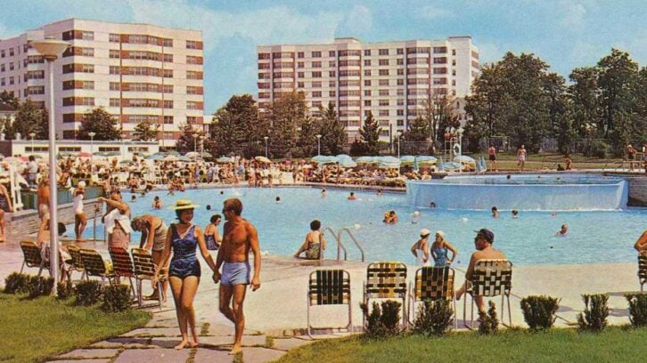 People relax and swim in a busy outdoor pool surrounded by grass and modern apartment buildings. Groups lounge on chairs while children and adults enjoy the water under a sunny sky.
