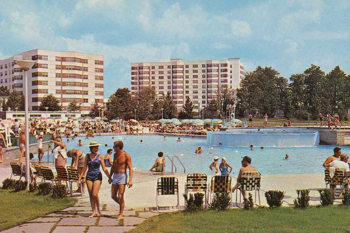 People relax and swim at a large outdoor pool surrounded by lounge chairs, with mid-century apartment buildings and trees in the background under a blue sky.