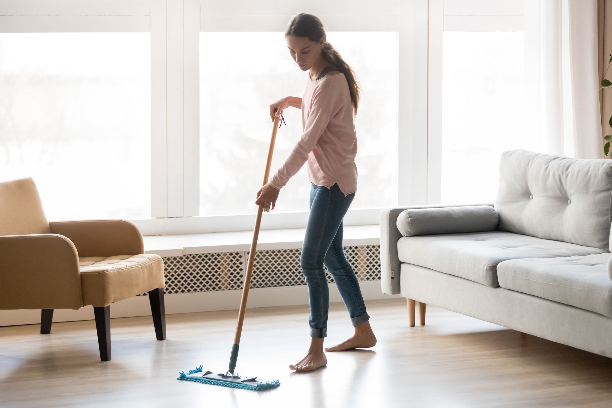 Full-length image of barefoot young woman stands in living room homeowner doing house chores cleaning wooden laminate floor using microfiber wet mop pad, housekeeping job or routine home work concept