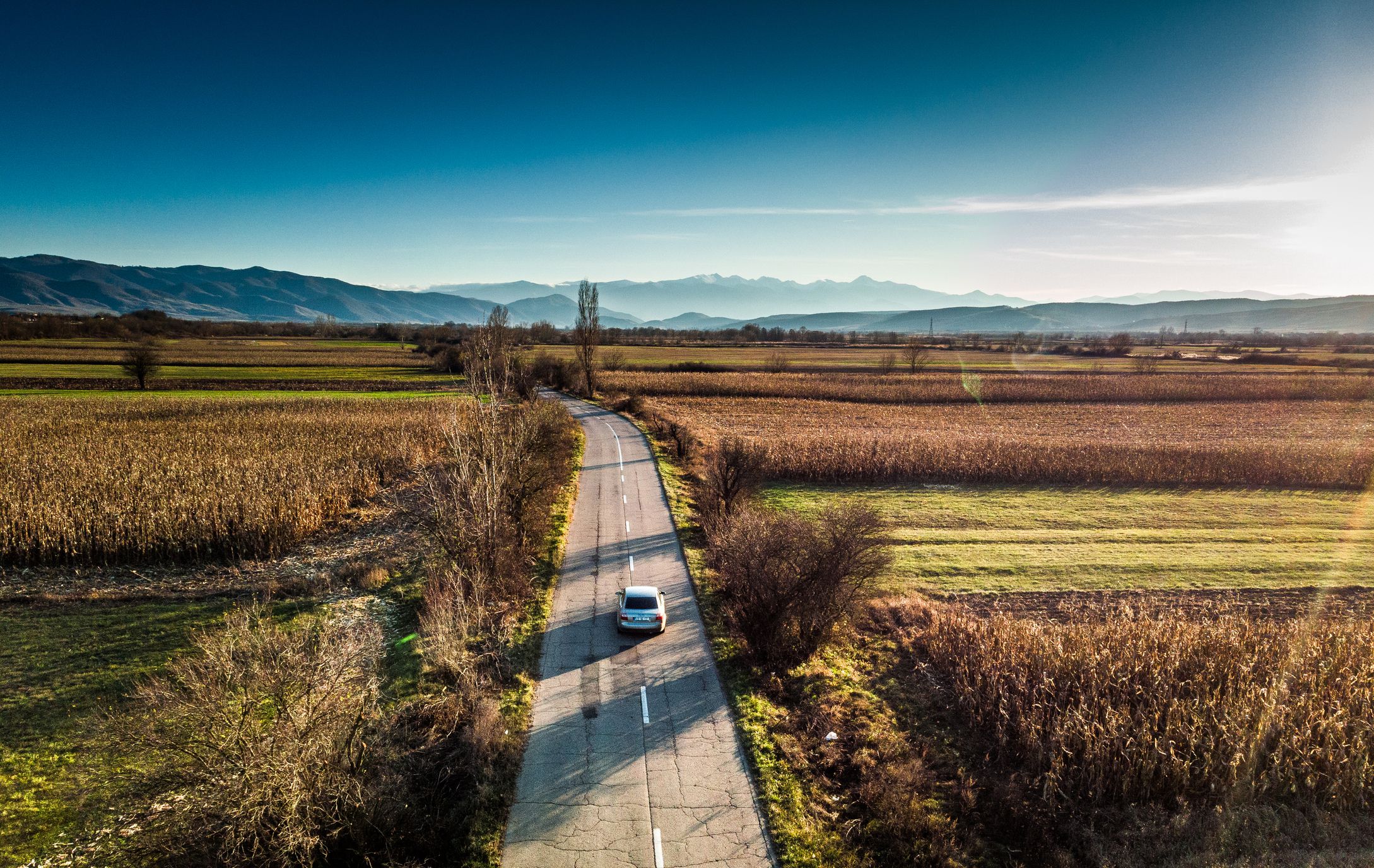 Color image depicting an aerial shot, taken by drone, of a car driving on a straight road flanked by lush agricultural meadows, corn fields and farm land. There is a mountain range on the horizon in the distance. Room for copy space.
