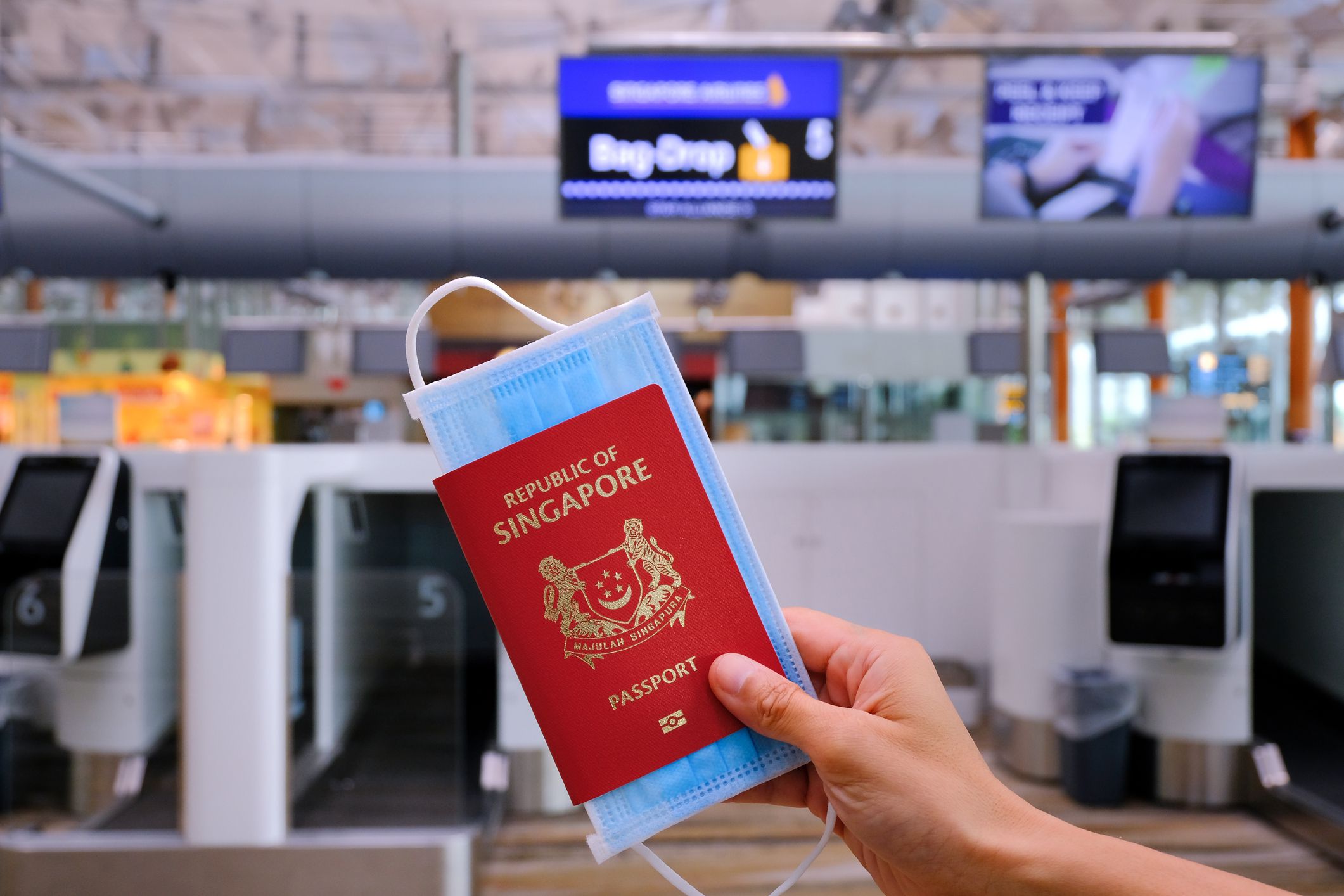 Airport check-in hall. Hand holding Singapore passport and face mask, at luggage check-in (bag drop). Travel concept. Reopening; coronavirus covid-19; travel restrictions.