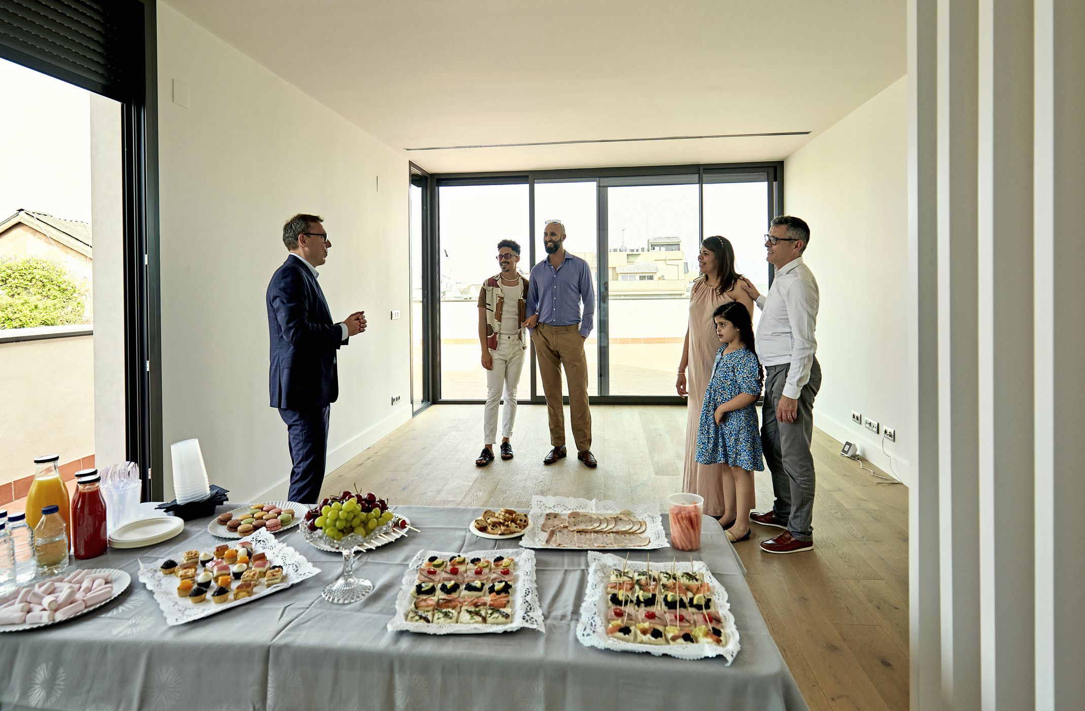 View across refreshment table of male realtor making sales pitch to family with one child and gay couple as they stand together in unfurnished apartment.