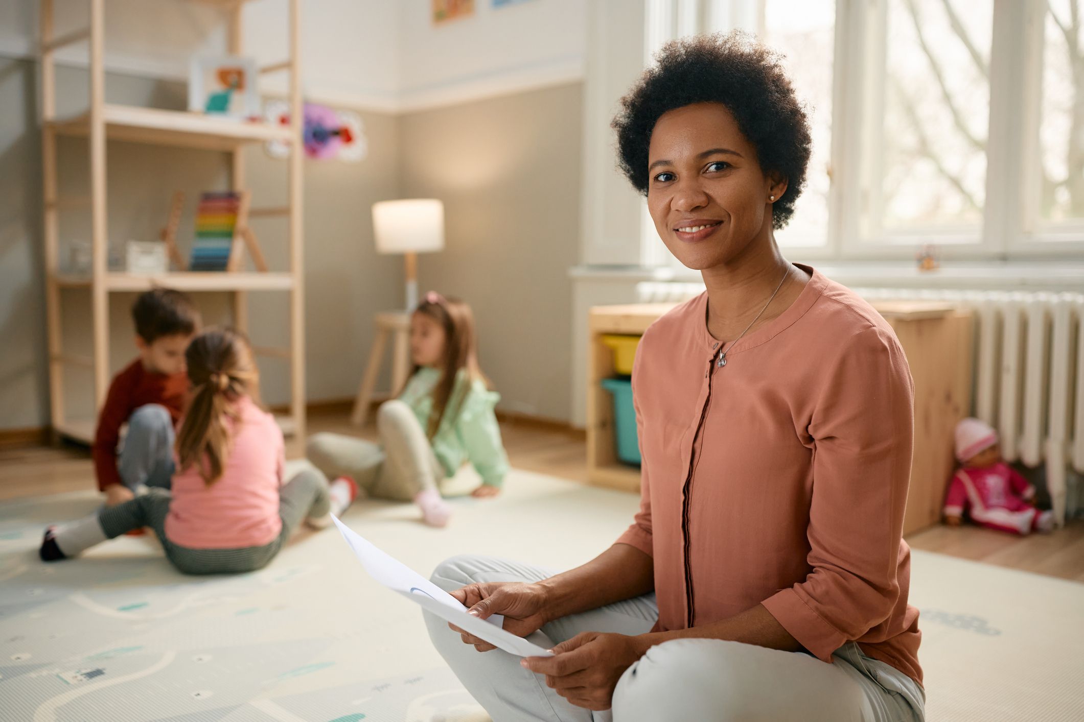 Smiling African American preschool teacher sitting on the floor and looking at camera. There are kids playing in the background.