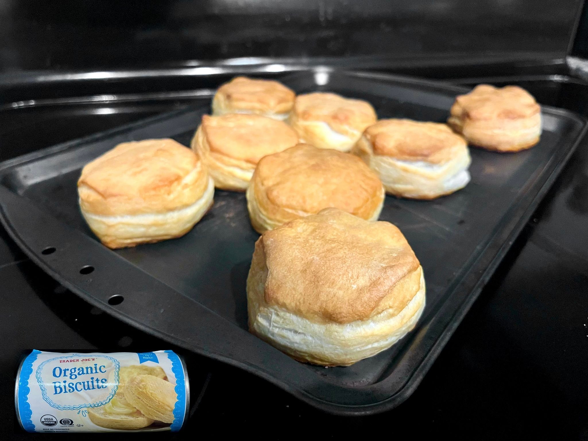 Eight golden-brown biscuits are on a baking tray. In the corner, a tube labeled "Organic Biscuits" shows an image of baked biscuits. The background is a black stovetop.