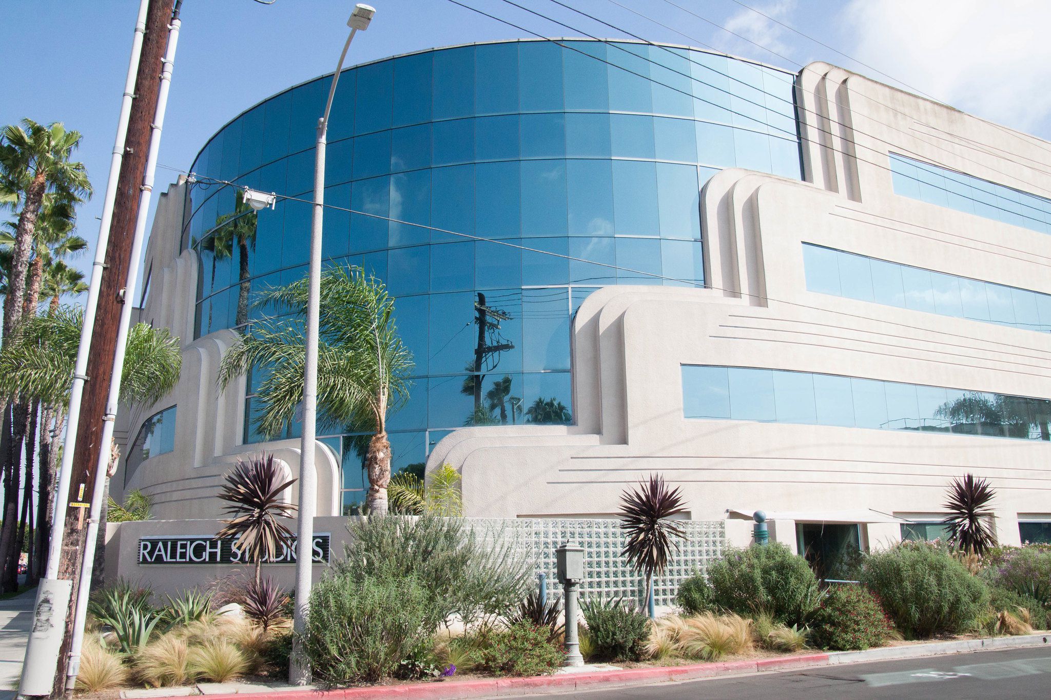 A modern building with curved glass windows and white concrete details, surrounded by palm trees and greenery. A sign in front reads "Raleigh Studios." Blue sky is visible in the background.