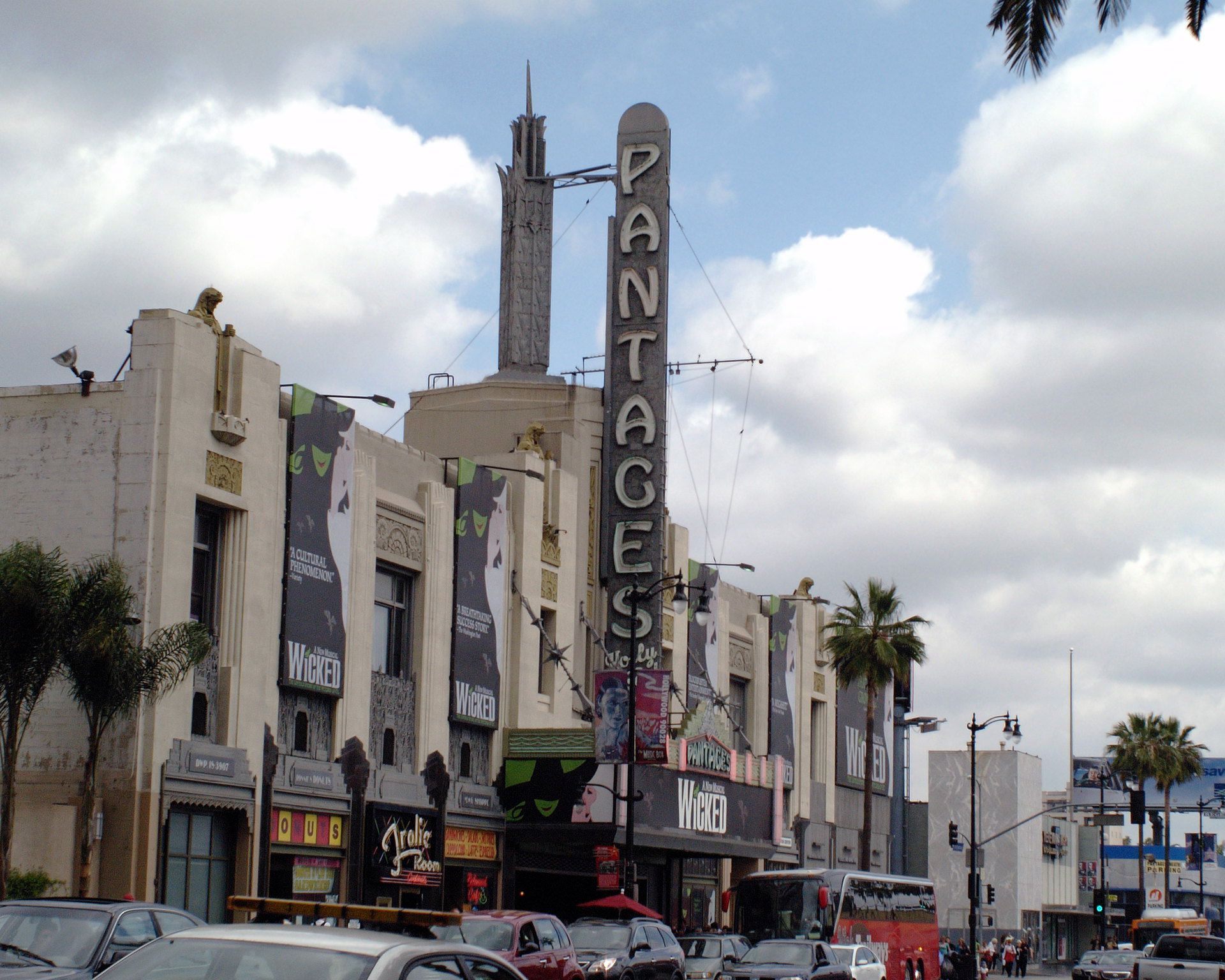 A large vertical Pantages Theater sign on a historic building in Los Angeles, with "Wicked" musical banners, palm trees, cars, and a cloudy sky in the background.