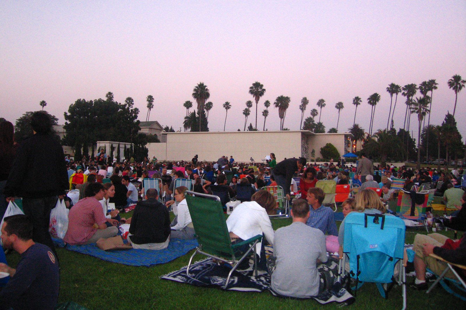 A large group of people sit on blankets and lawn chairs on a grassy field at dusk, enjoying an outdoor event with palm trees in the background.