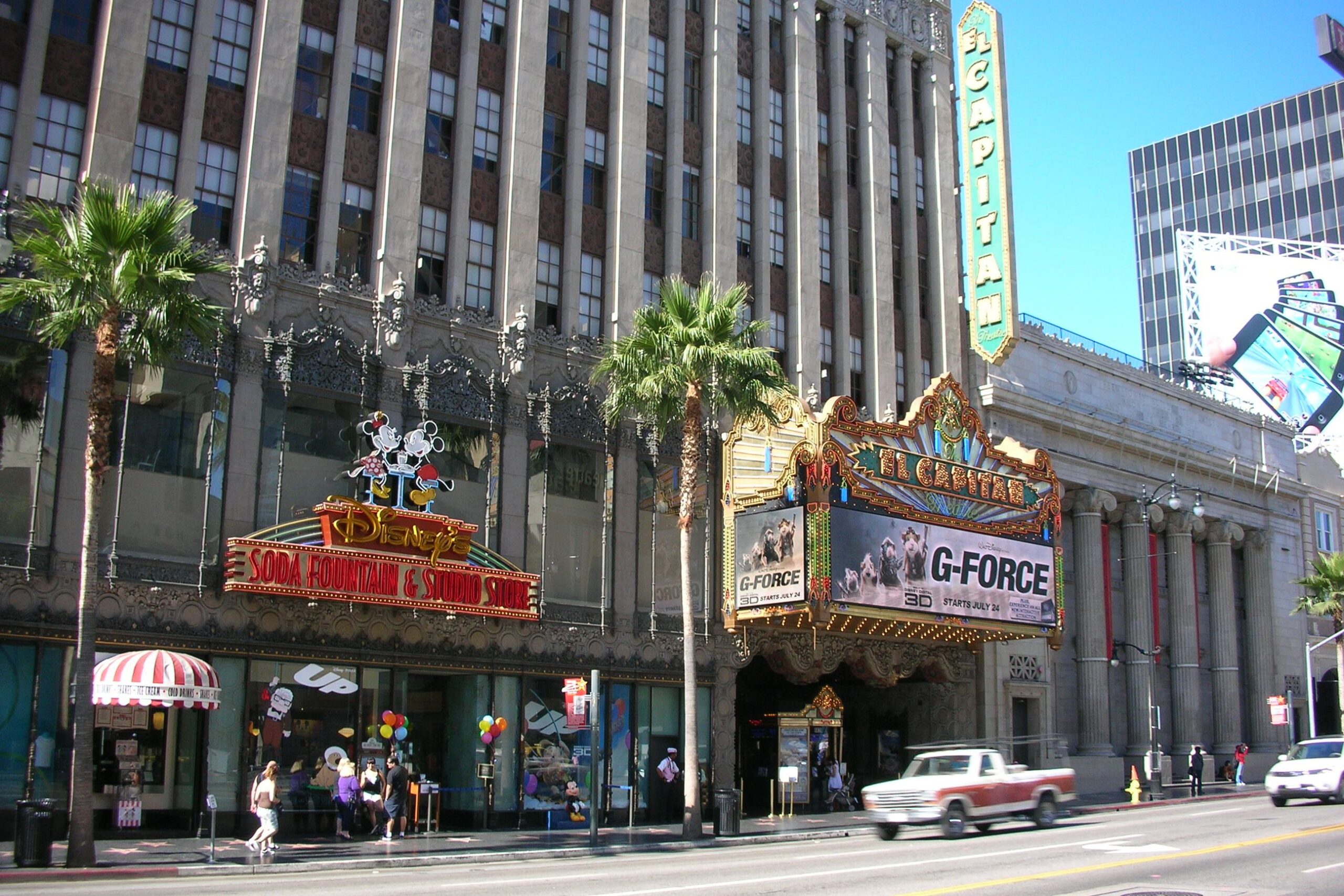 Street view of the El Capitan Theatre in Hollywood, featuring a marquee advertising “G-Force” and the Disney Soda Fountain & Studio Store entrance, with palm trees and cars passing by.